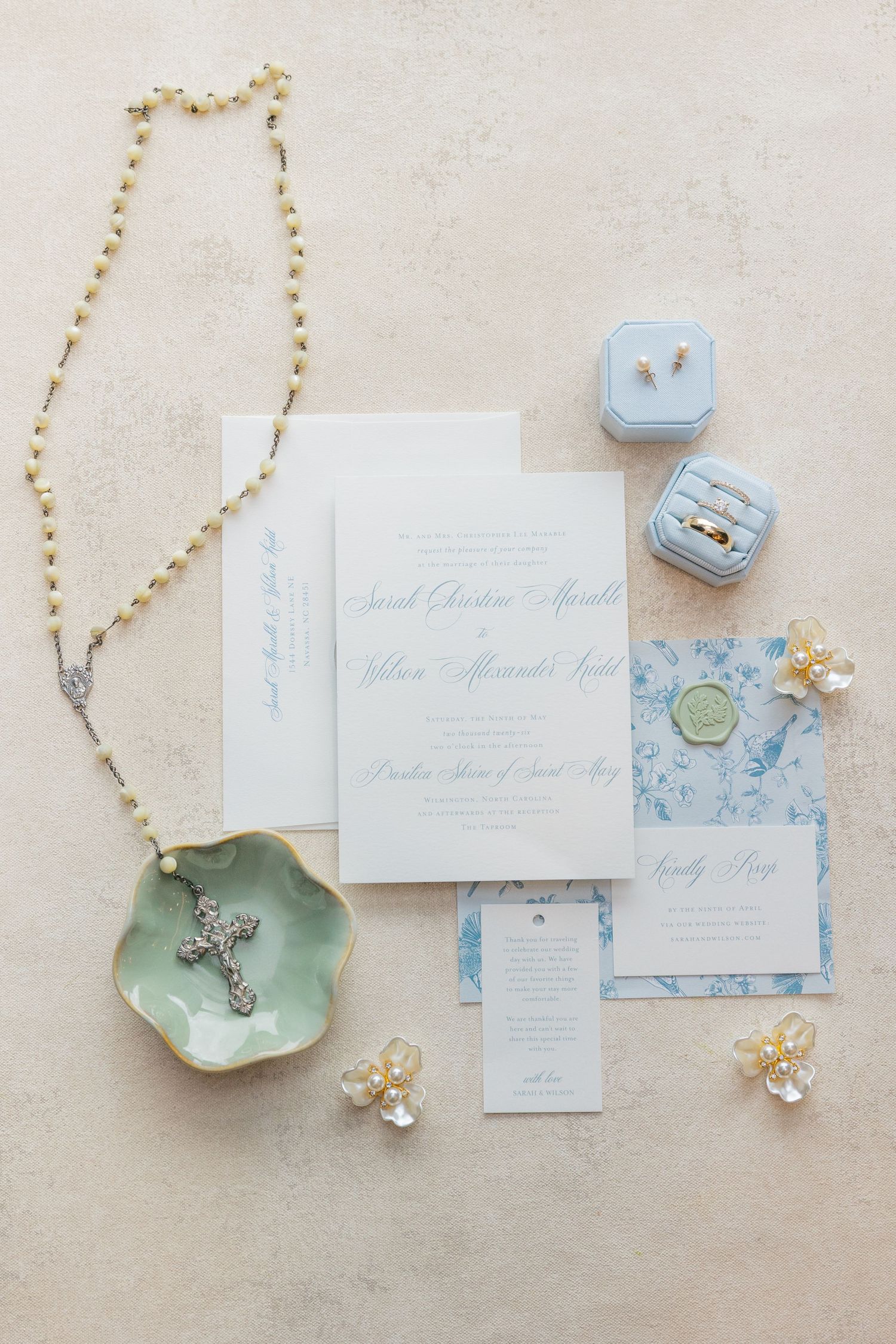 Elegant wedding flat lay with blue invitations, rings, rosary, and pearl jewelry on cream background.