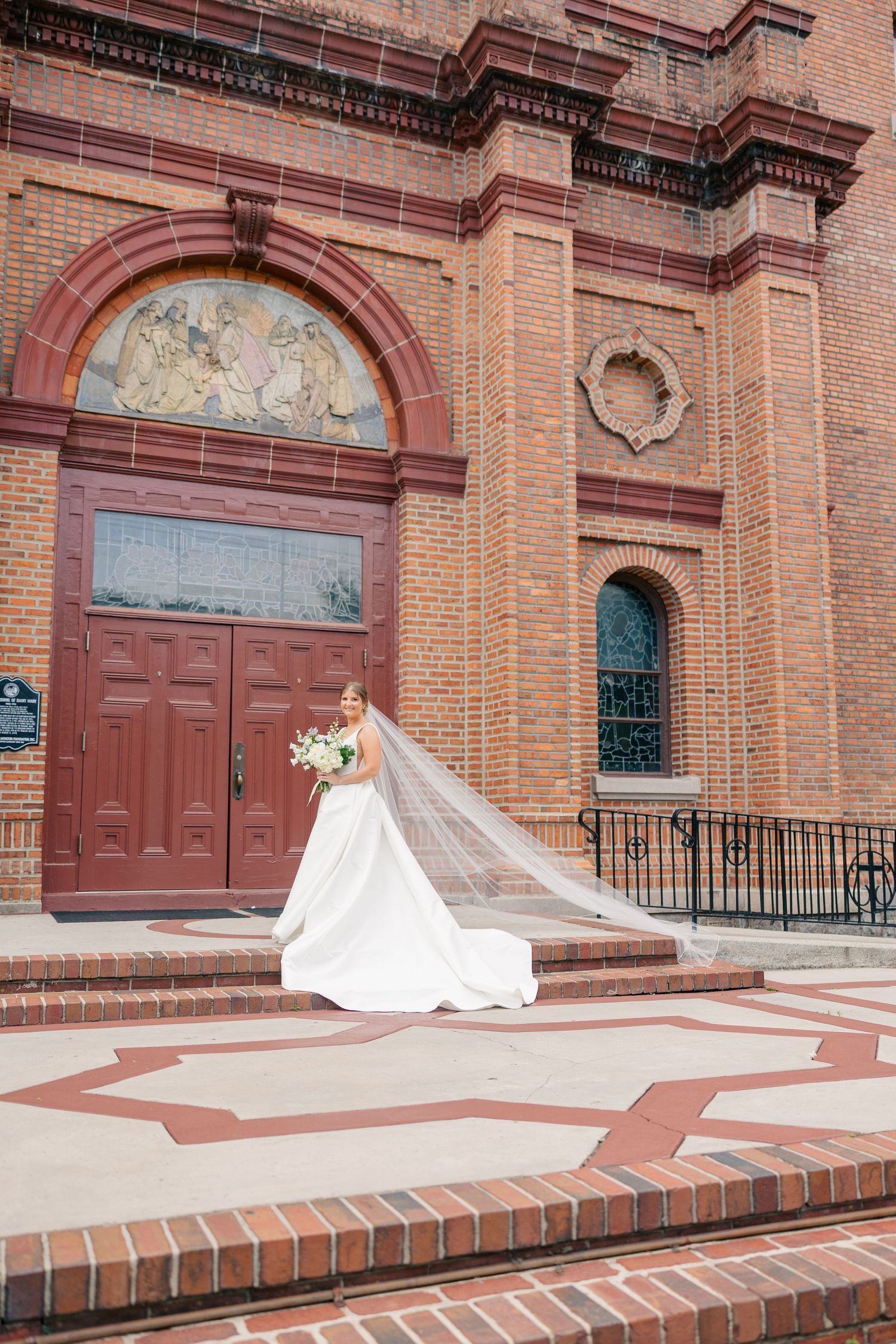 Bride in a white ballgown with a long flowing veil stands on the steps of a grand red brick church.