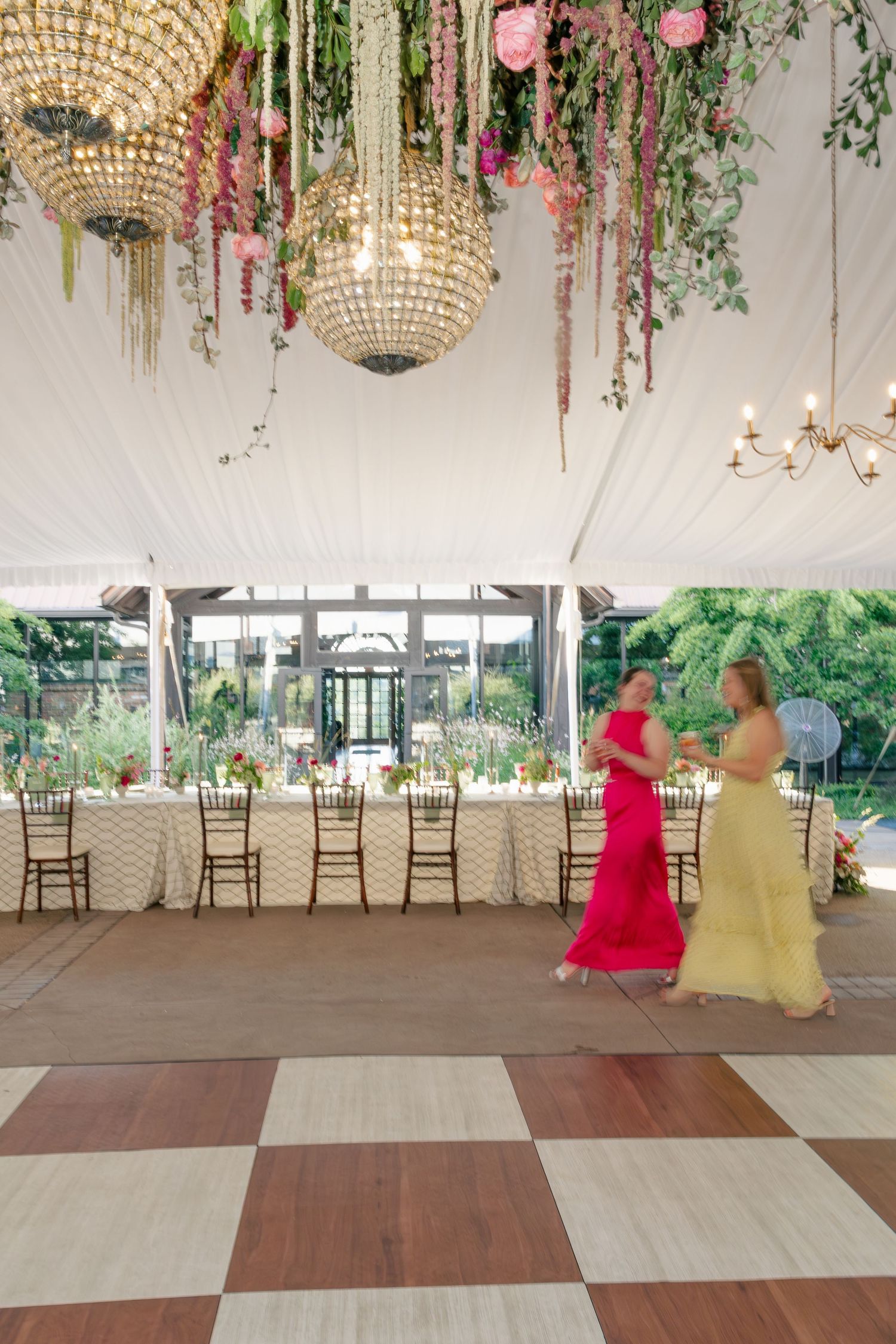 Two women in colorful gowns walk through elegant tented reception hall with floral chandelier decor.