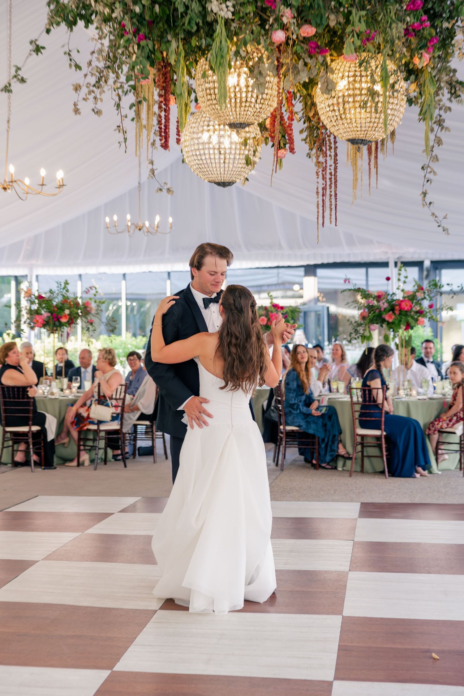 Bride and groom share first dance under crystal chandeliers adorned with cascading florals in tent.