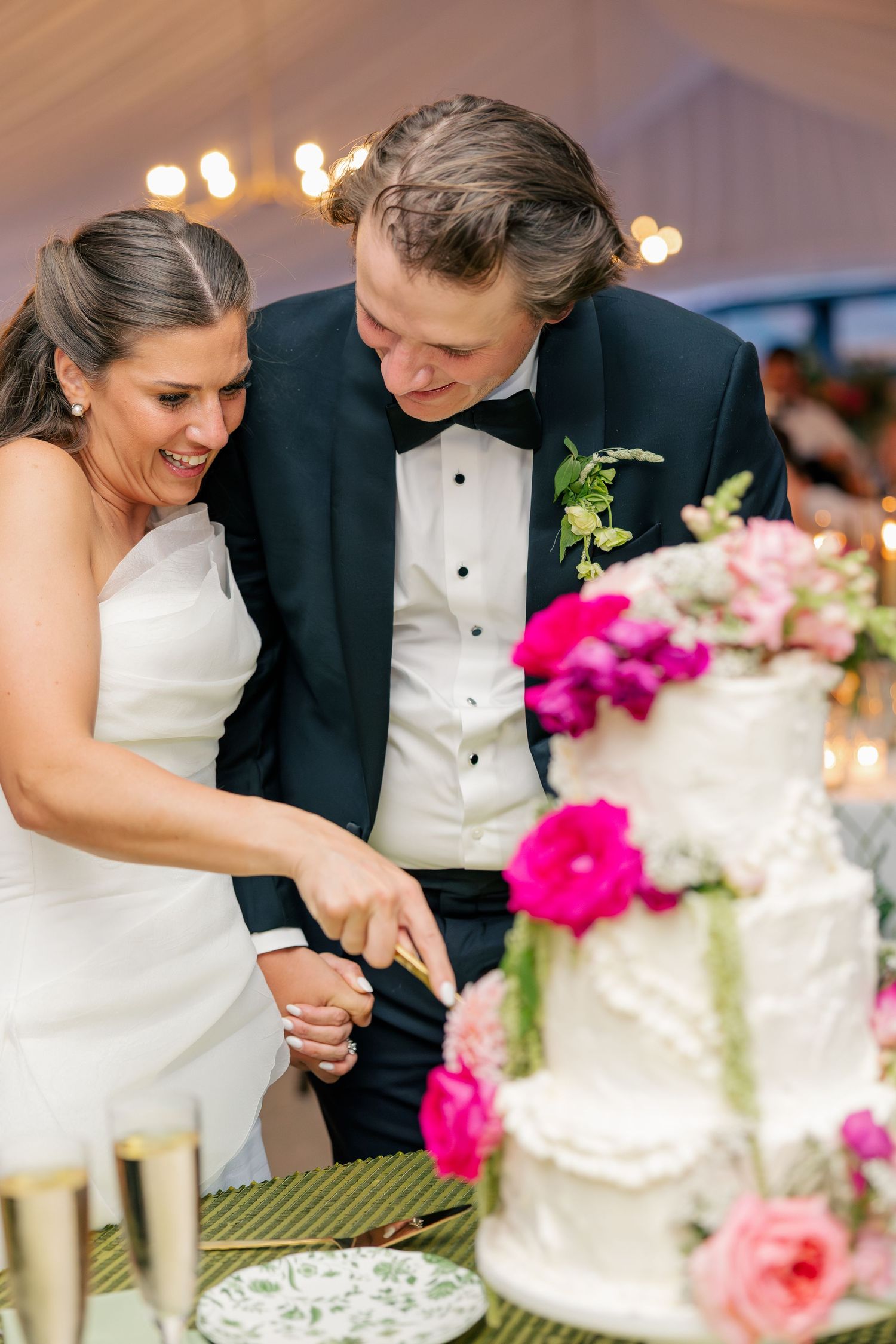 Bride and groom smile while cutting their white three-tier wedding cake adorned with pink flowers.