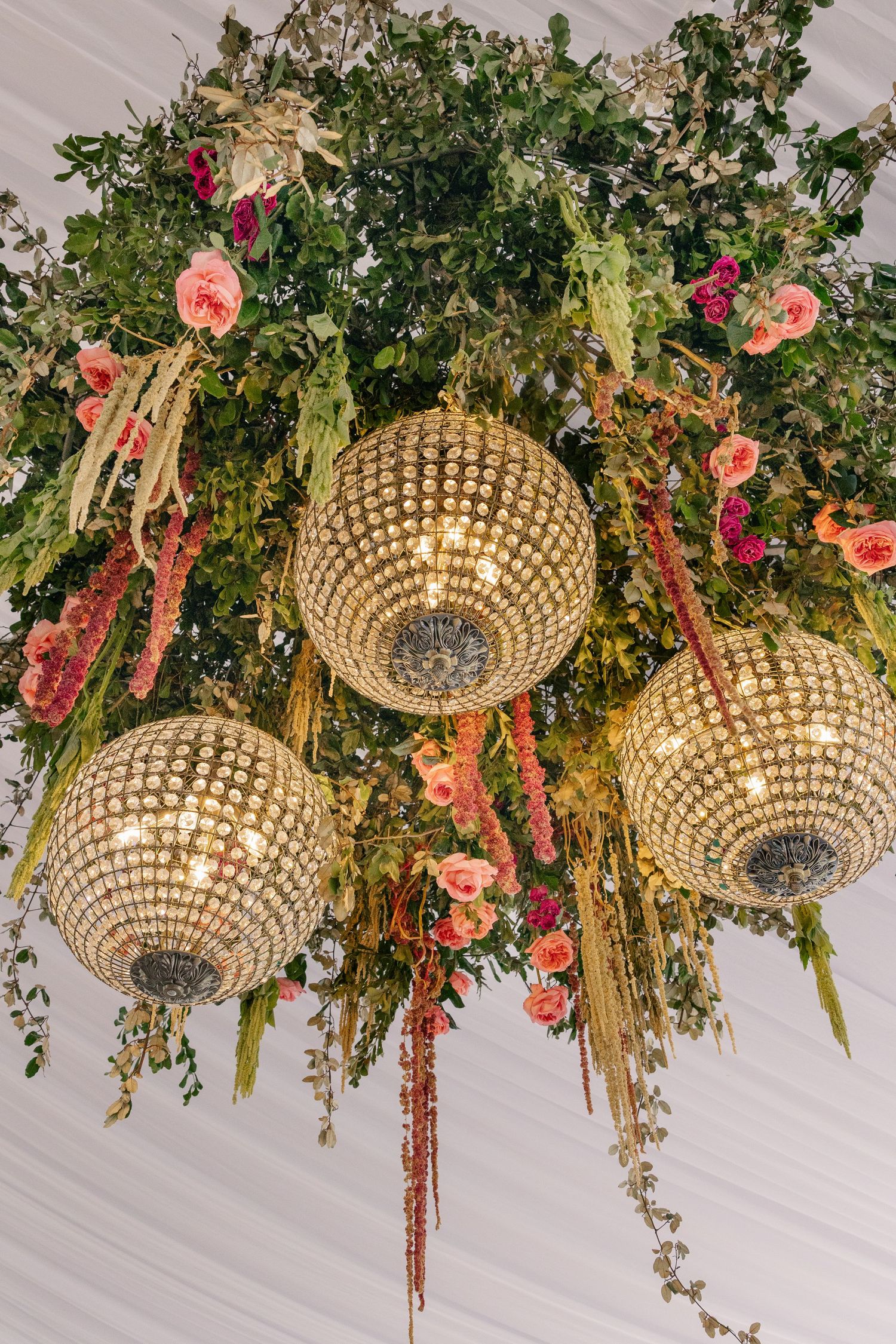 Glamorous crystal globe chandeliers surrounded by lush greenery and pink floral arrangements hang from a tent ceiling.