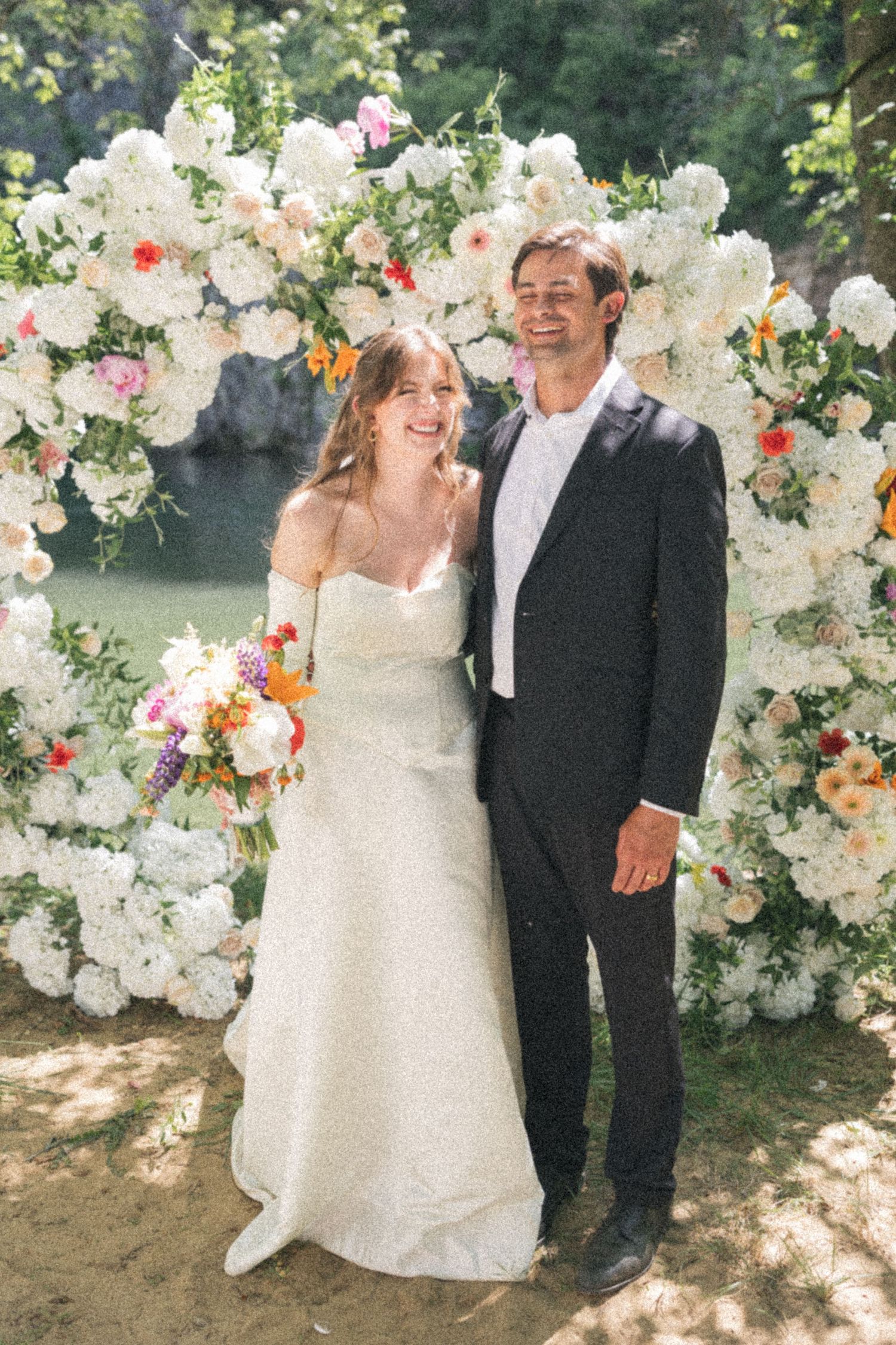 Smiling bride and groom posing in front of a lush white floral arch with colorful accents outdoors.