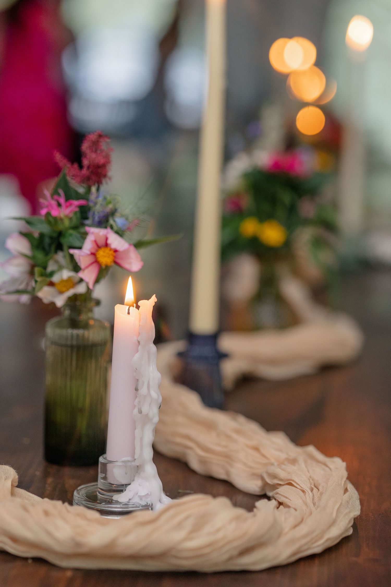 Lit white pillar candle on a rustic wooden table with wildflowers in green glass vases at a wedding reception.