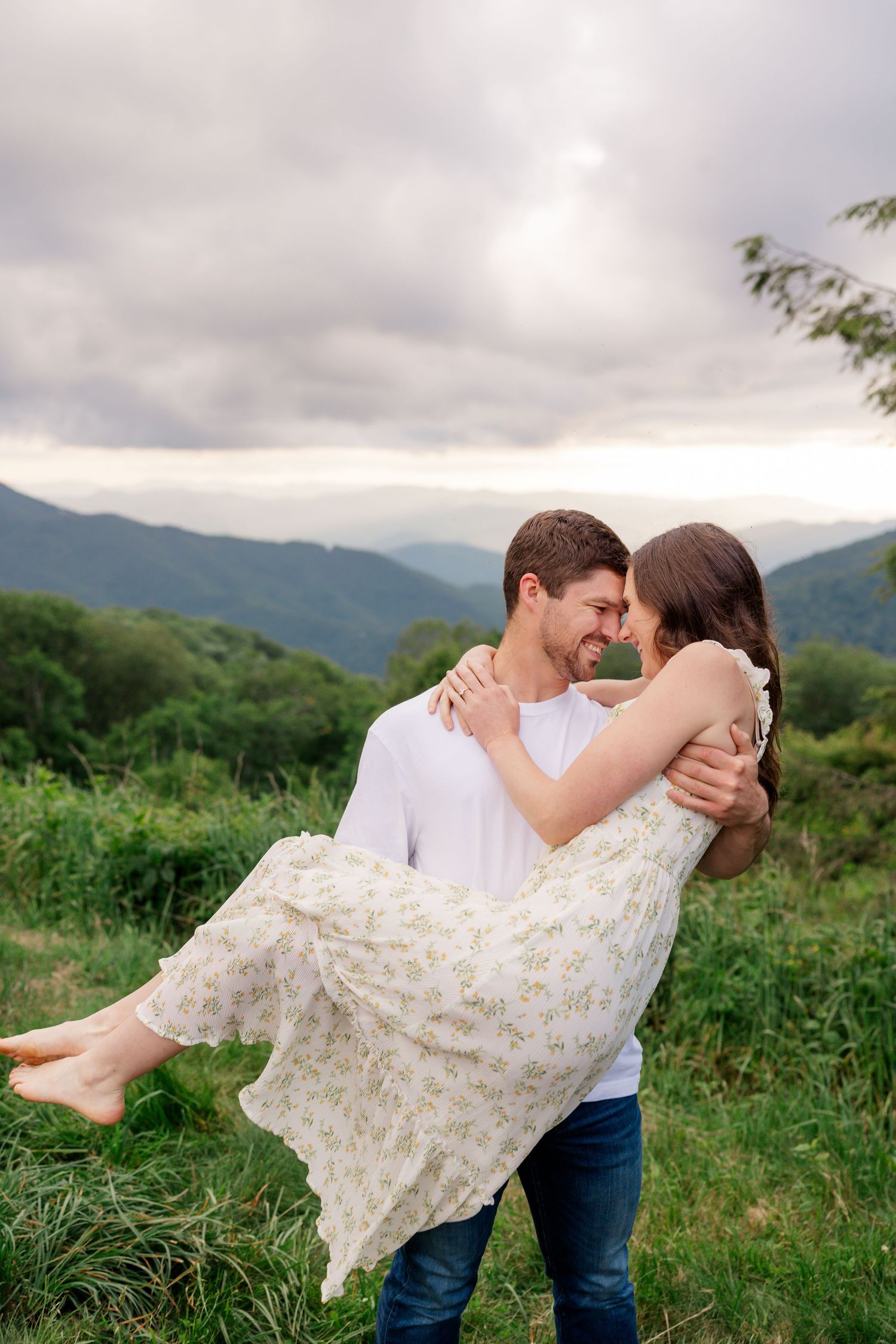 A couple shares romantic moments in a mountain meadow as the man lifts the woman wearing a floral dress.