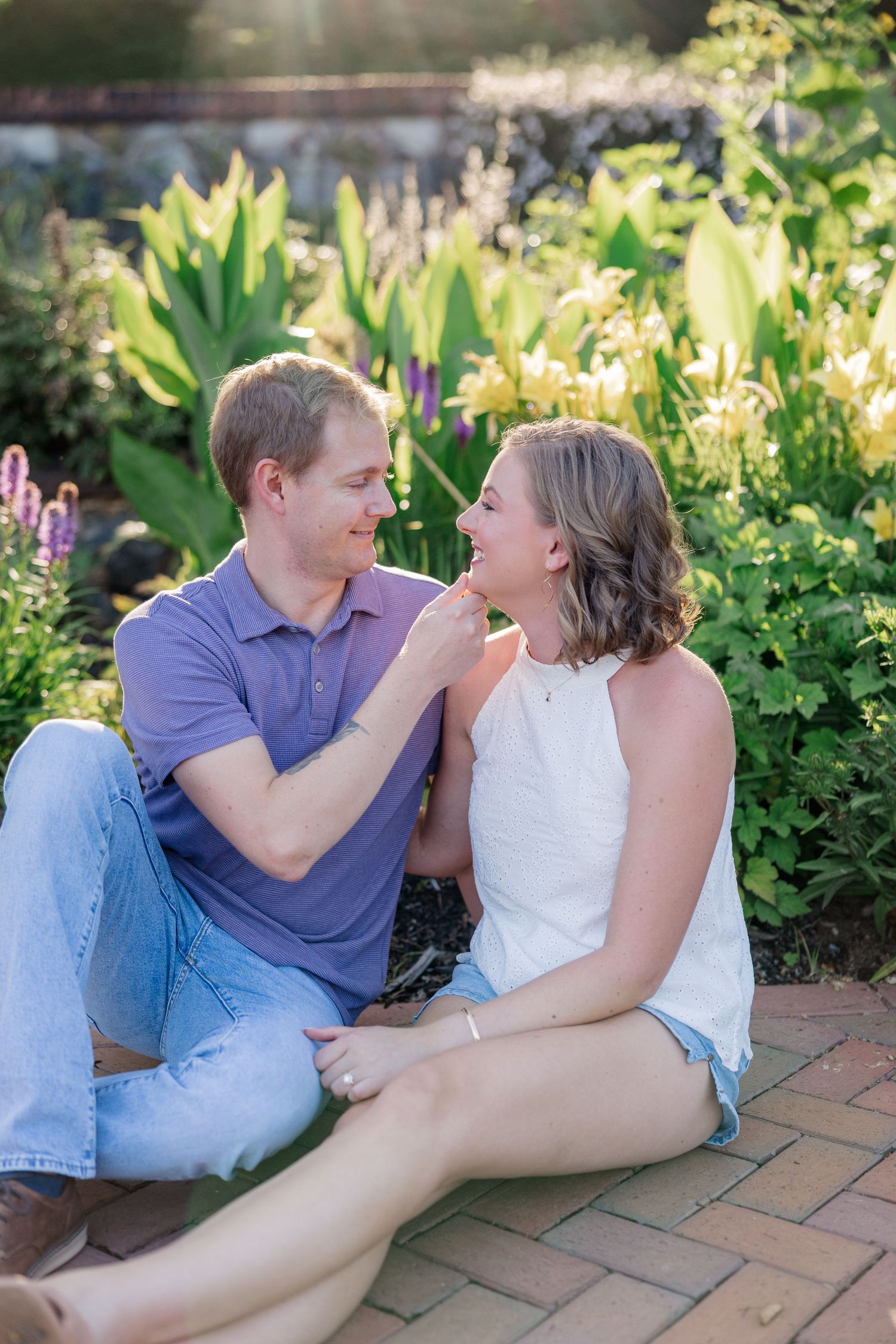 A couple shares playful moments while relaxing on brick pavers in a sunlit garden with blooming flowers.