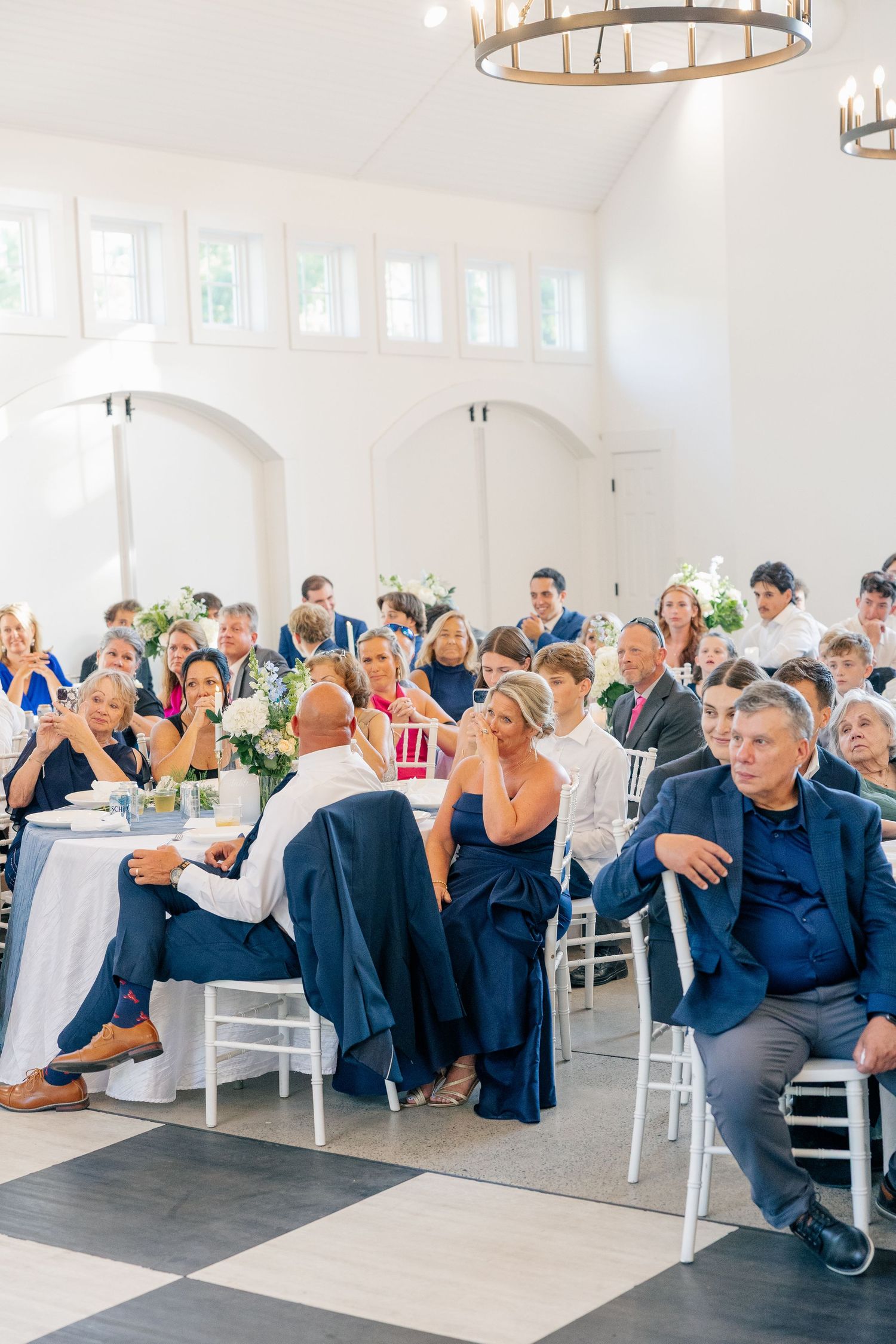 Wedding reception guests seated at tables in a bright white ballroom during speeches.