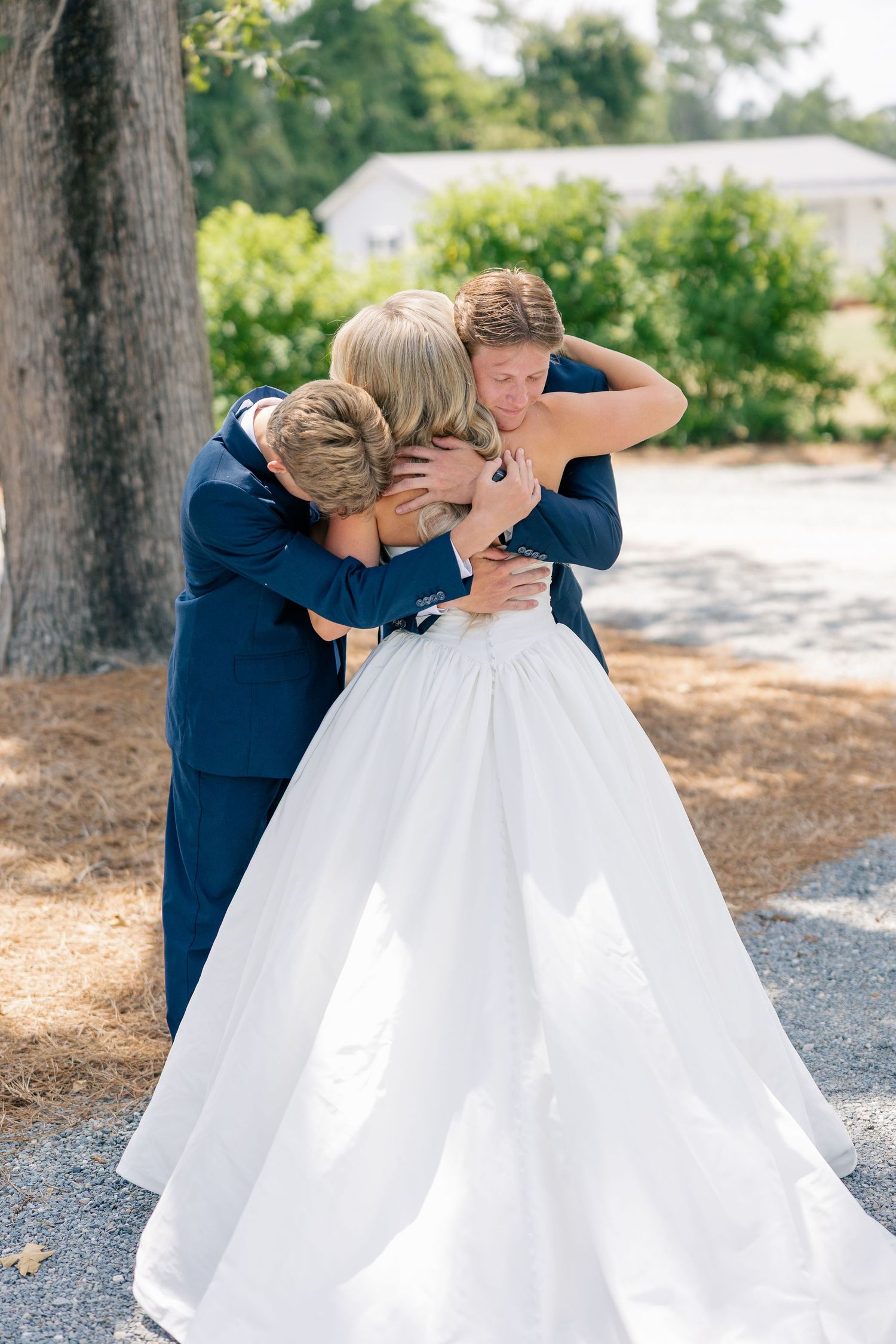 Emotional wedding moment as bride and groom share a group hug with a family member outdoors.