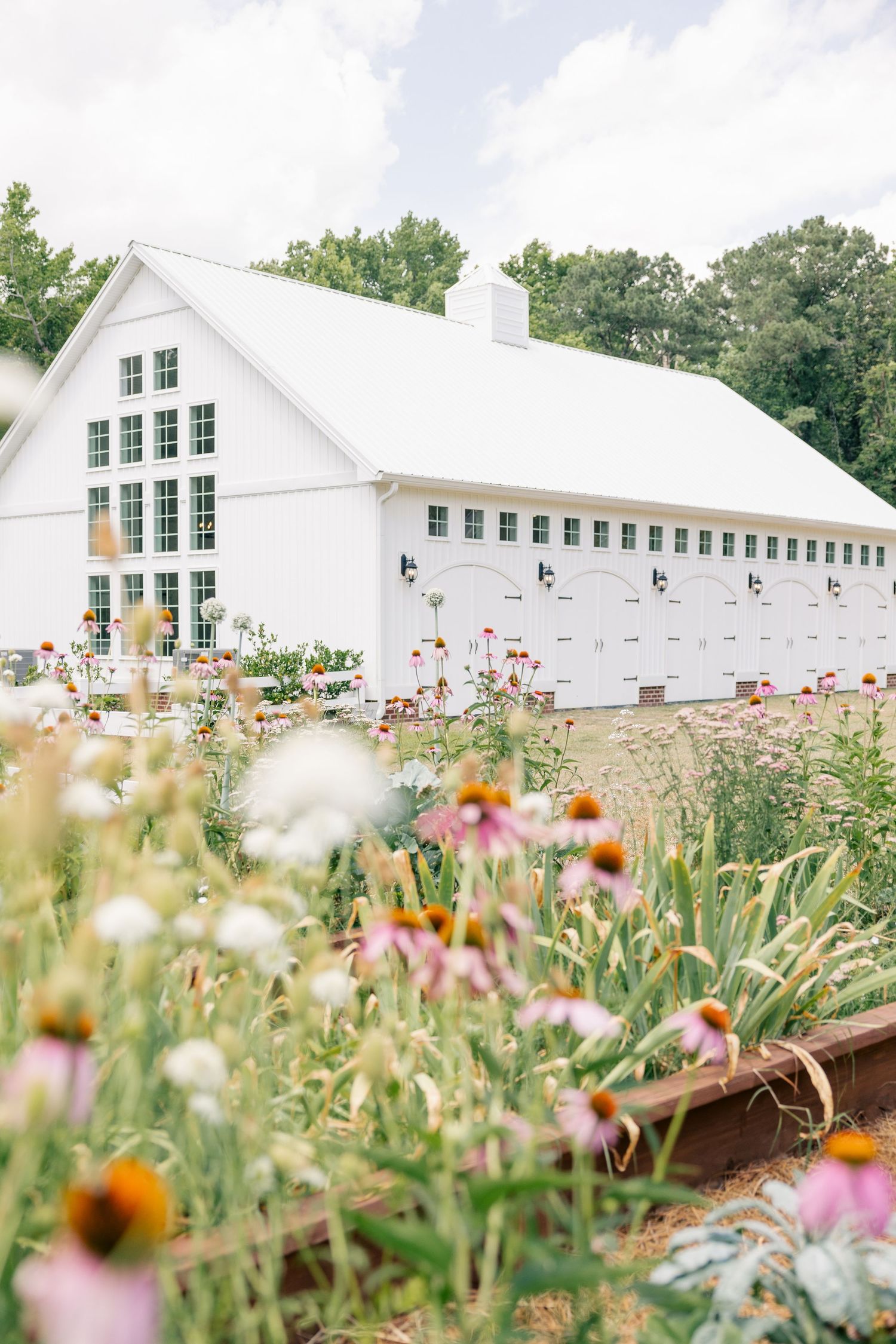 White barn wedding venue surrounded by colorful wildflower gardens under a partly cloudy sky.