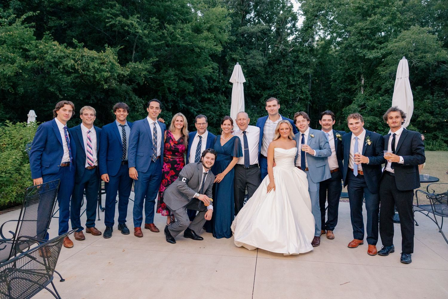 Wedding party group photo outdoors, bride in white gown surrounded by groomsmen and guests in navy and grey suits.