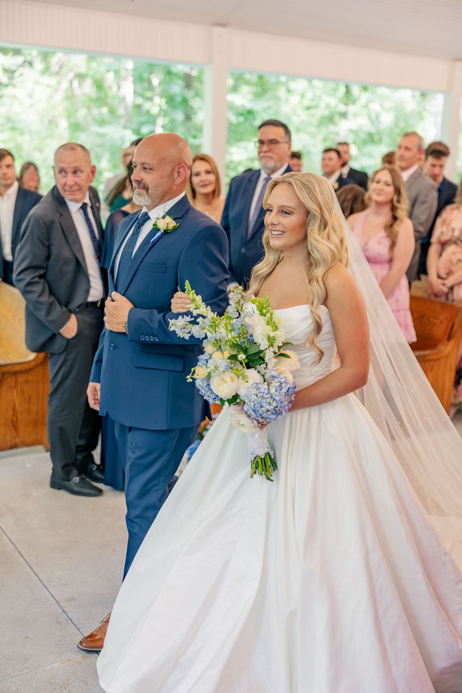 Bride walking down aisle with father, holding blue and white bouquet in sunlit outdoor chapel ceremony.