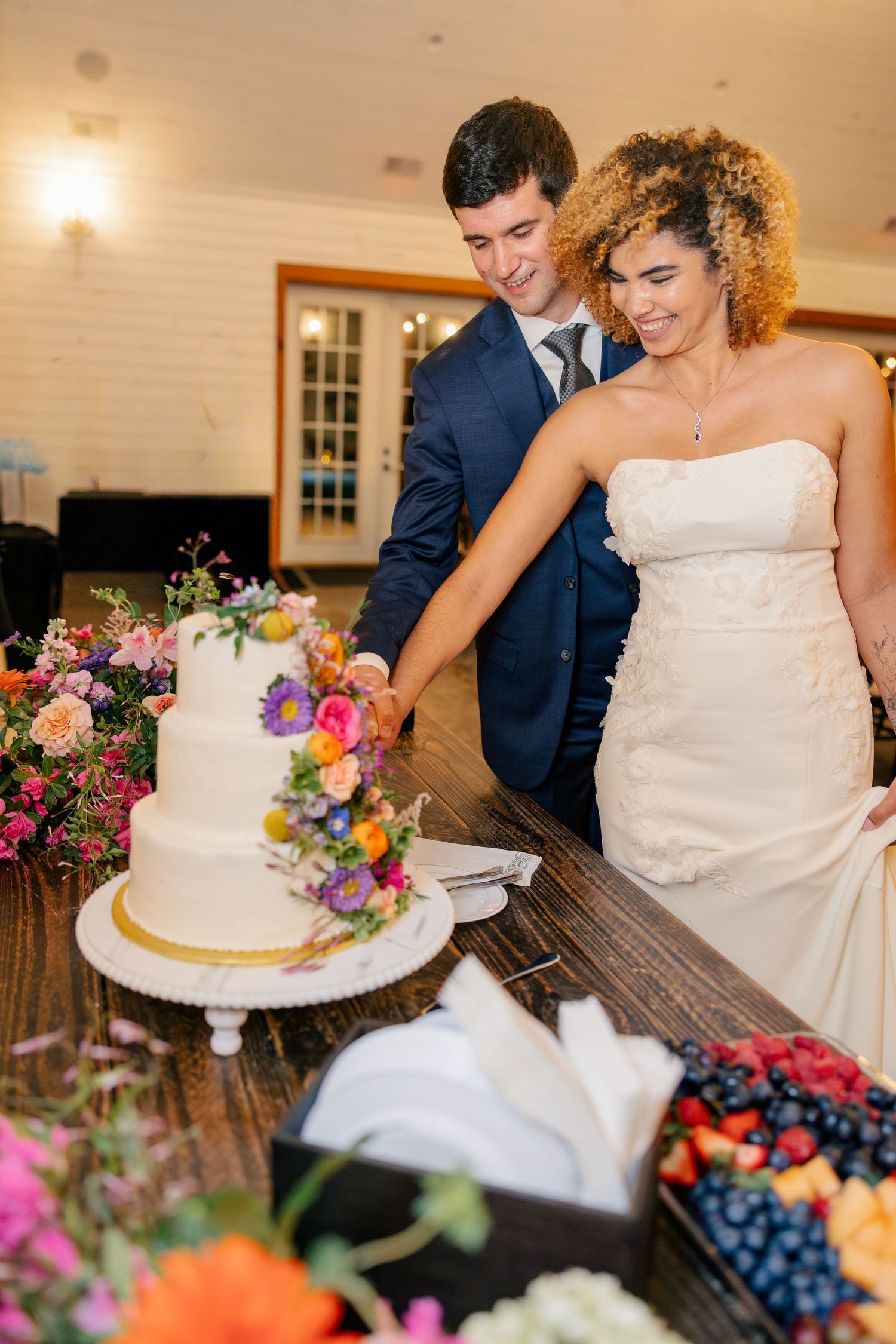 Bride and groom cut three-tier white wedding cake decorated with colorful flowers at reception.