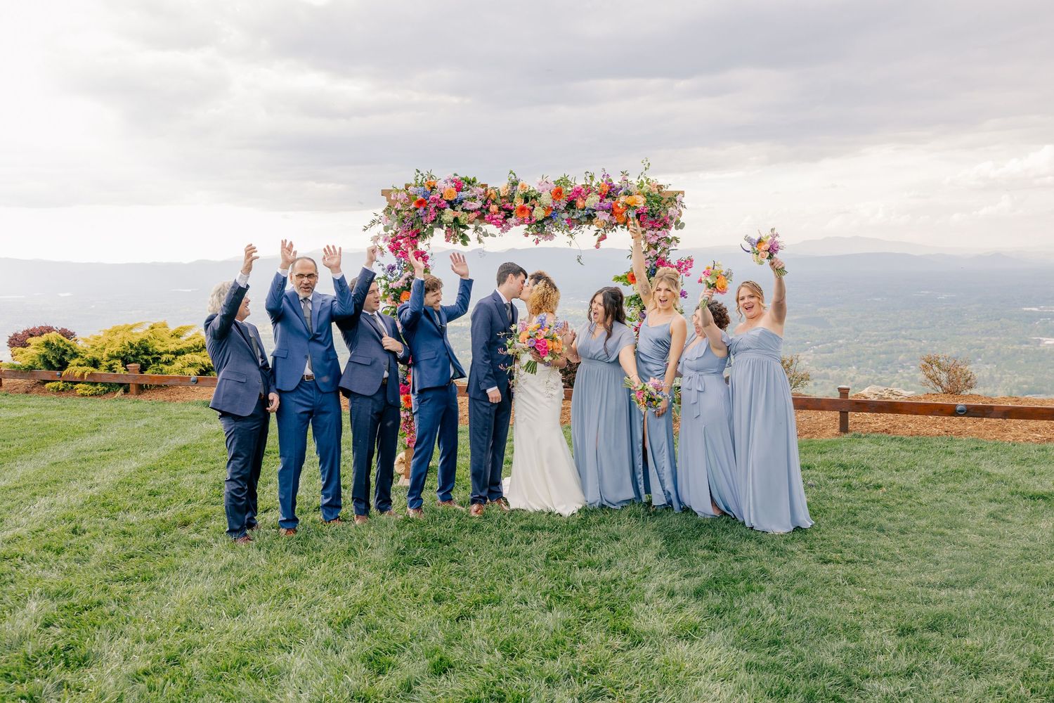 Wedding party celebrating under colorful floral arch with mountain views in background.