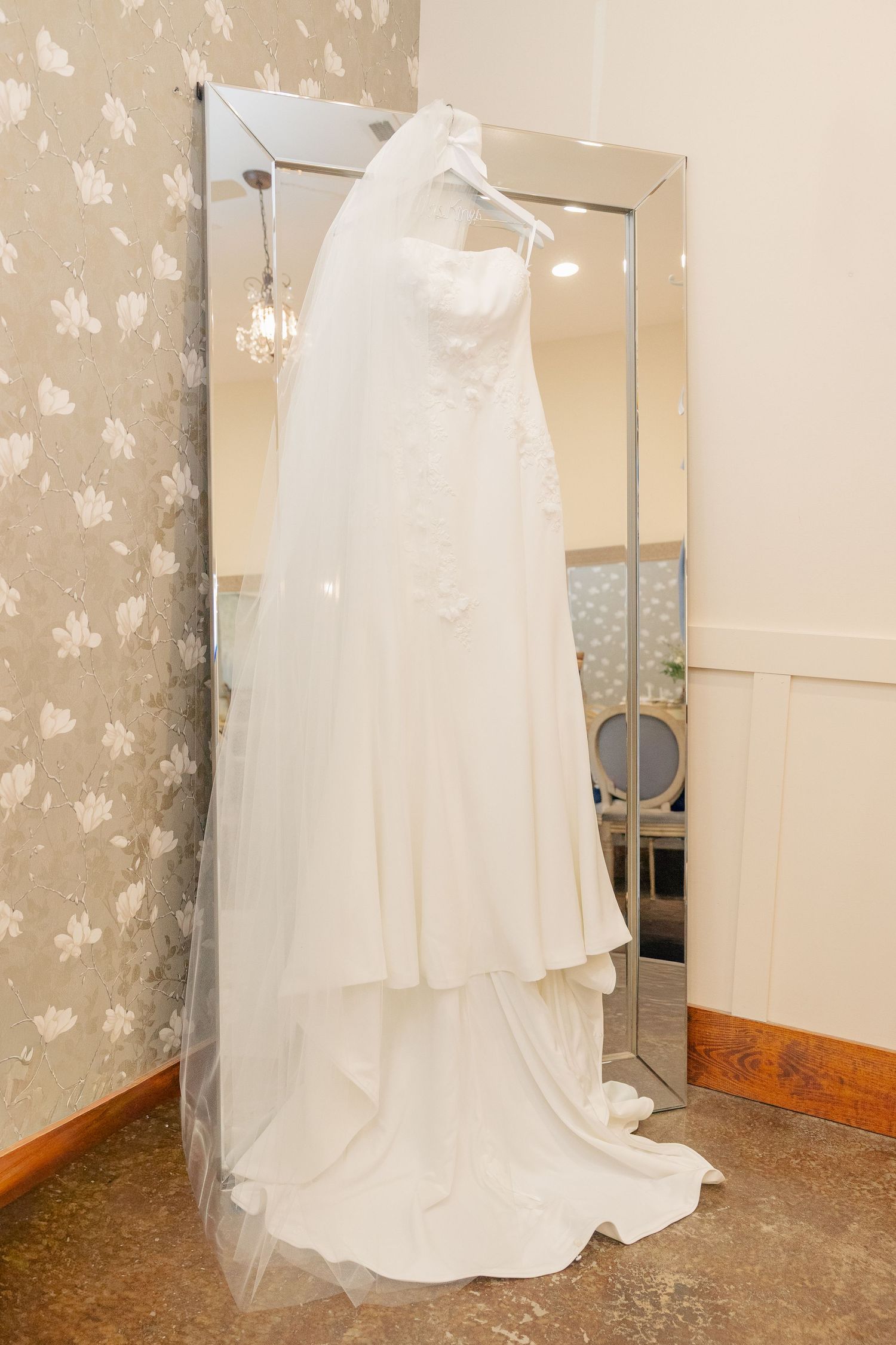 White wedding dress with veil hanging on a mirror in an elegant bridal suite with floral wallpaper.