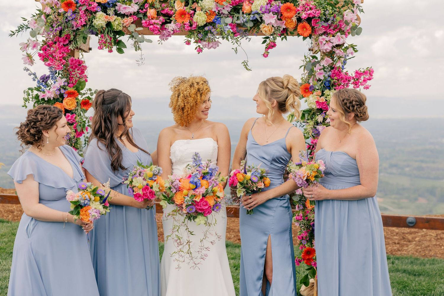 Bride and bridesmaids in blue gowns pose under vibrant floral arch with mountain valley views.