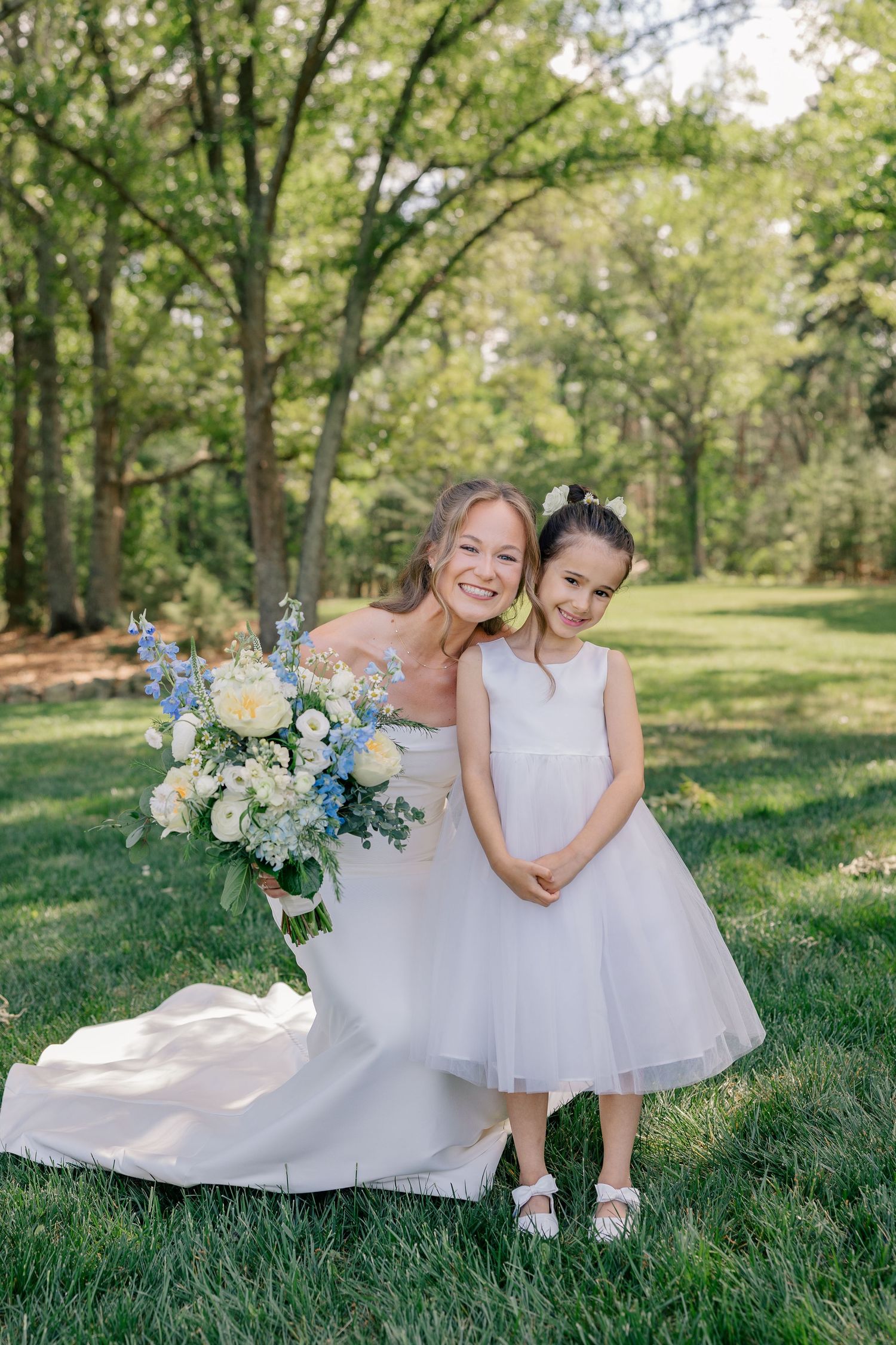 Bride in white gown holds blue and white bouquet while posing with young flower girl in white dress outdoors.