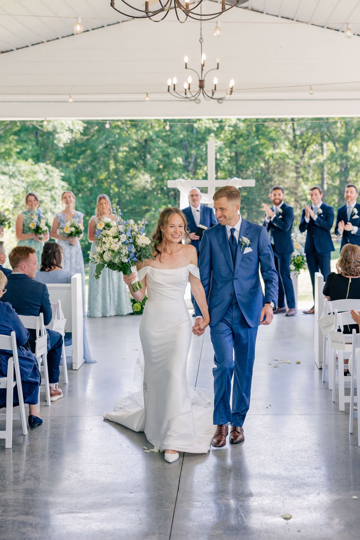 Happy bride and groom exit wedding ceremony holding hands in bright white tent venue with floral decor.