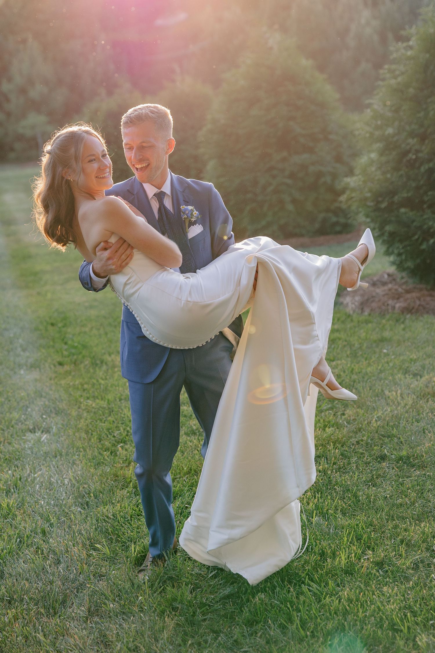Laughing groom carries smiling bride in elegant white gown in golden sunset light among evergreen trees.