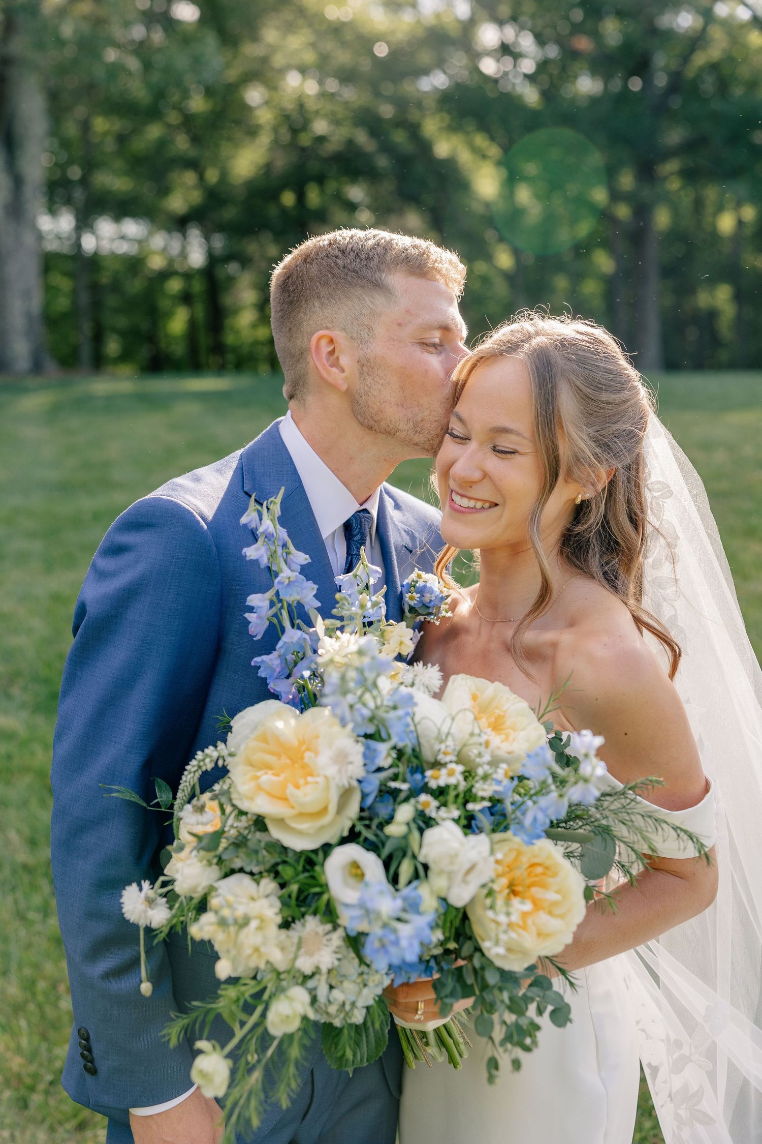 Groom in blue suit kisses smiling bride on cheek outdoors while she holds a yellow and blue bouquet.