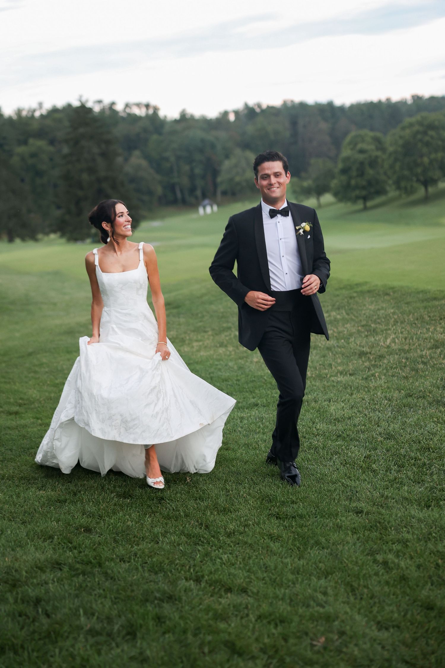 Bride in white gown and groom in black tuxedo walking on lush green grass at outdoor wedding venue.