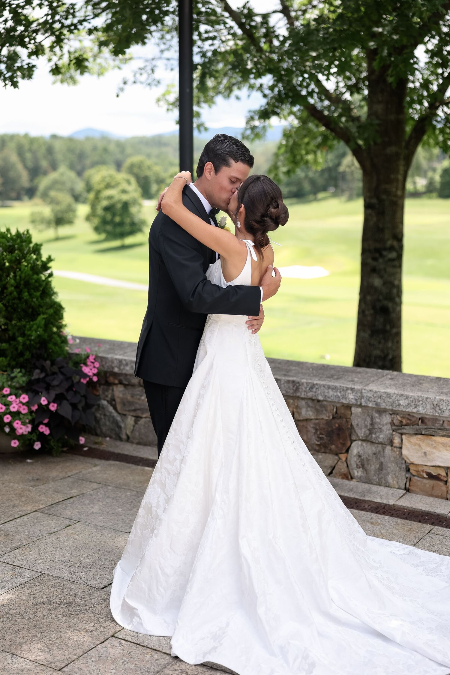 Bride and groom share a romantic first kiss in wedding attire on a stone terrace overlooking a lush green golf course.
