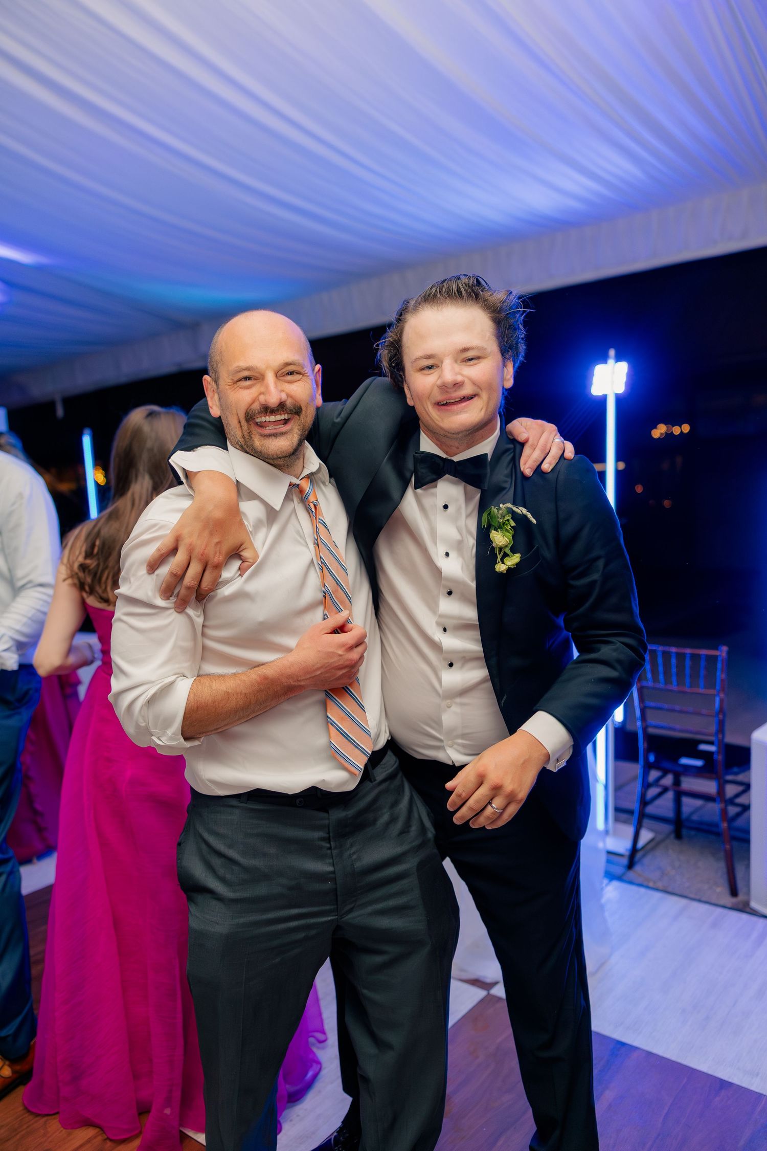Two men in formal attire smile and embrace on a wedding reception dance floor with blue lighting.