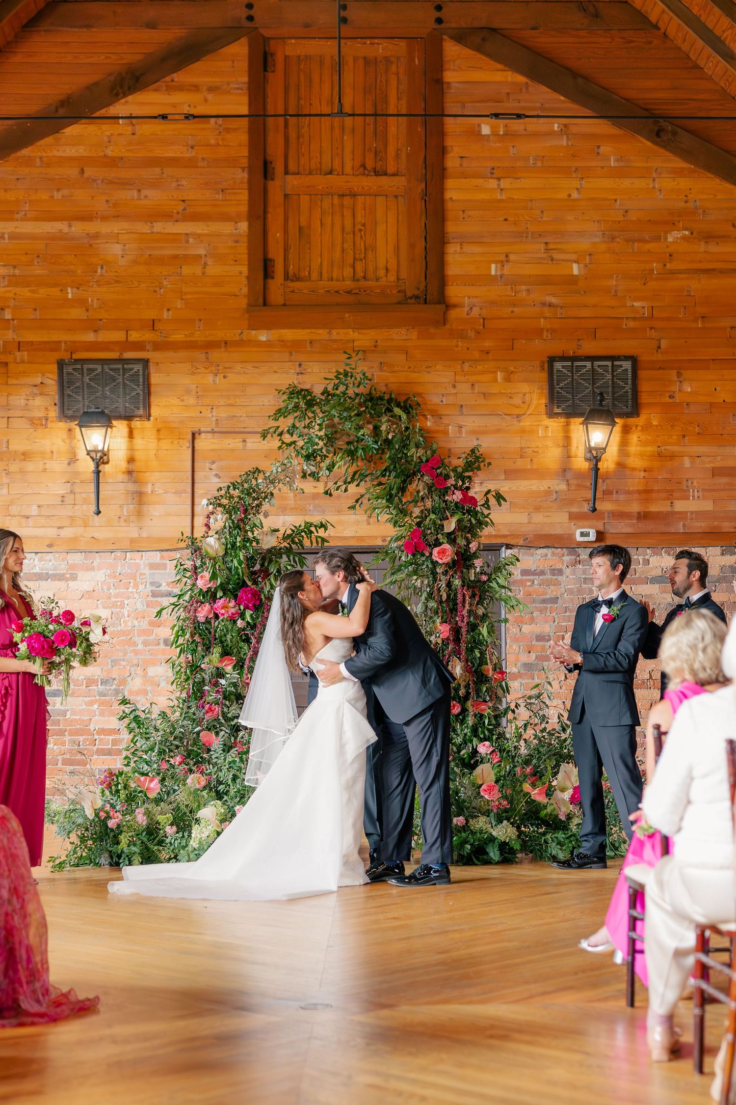 Bride and groom share first kiss under lush floral arch in warm wooden wedding venue.
