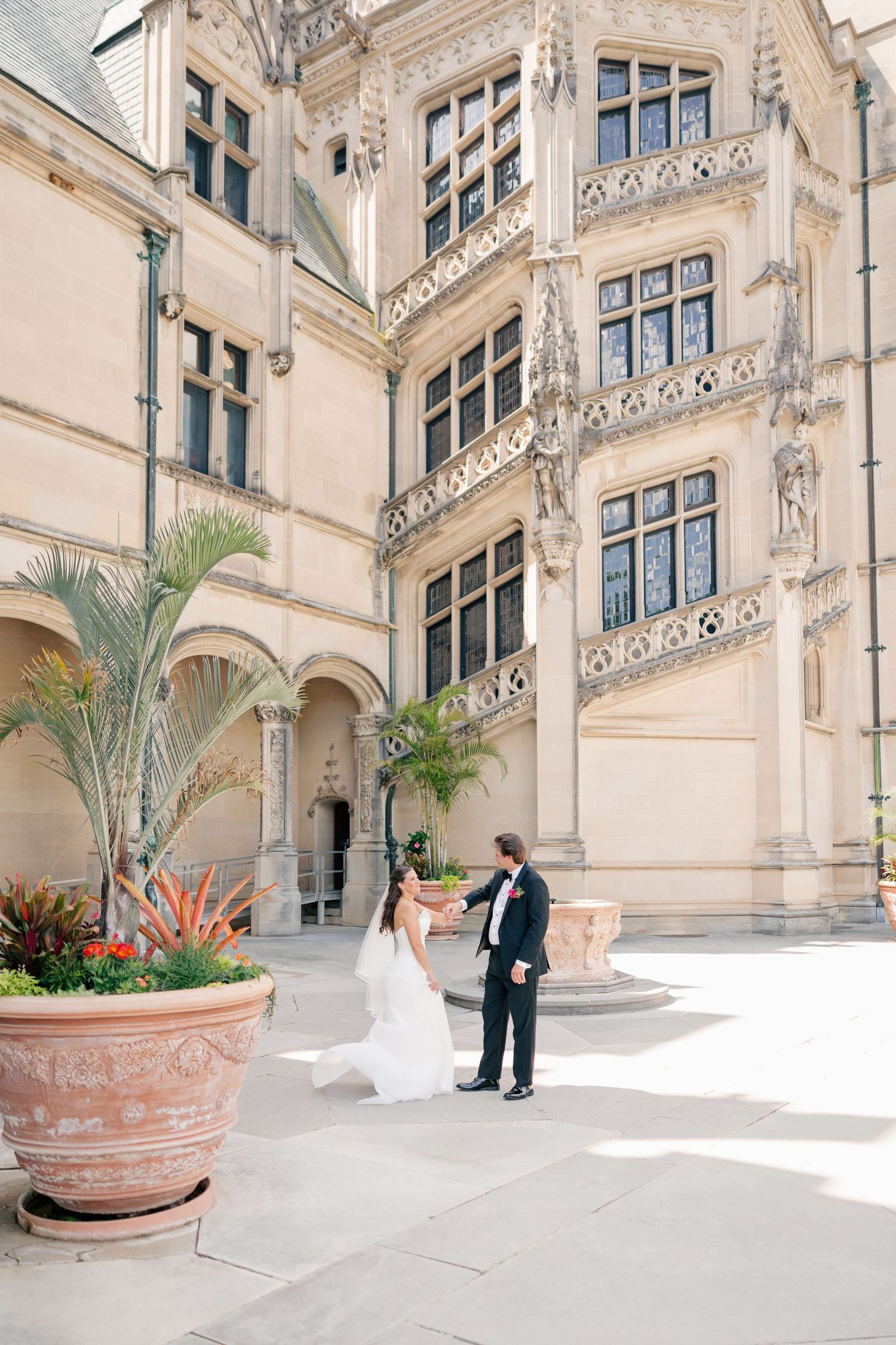 Bride and groom share first look in ornate castle courtyard with large tropical potted plants.