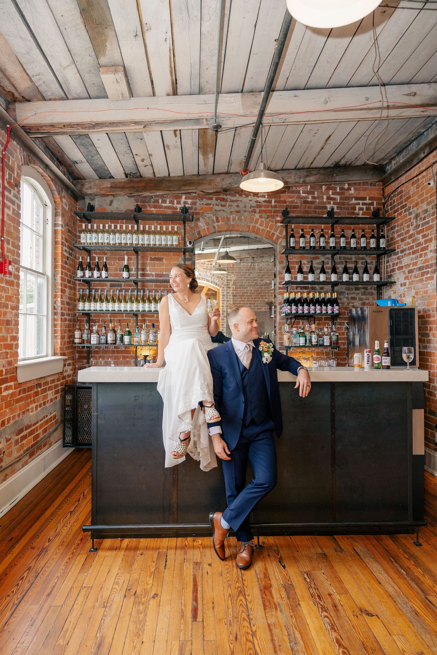 Bride sitting on bar counter with groom leaning beside her in rustic brick venue with wood floors.