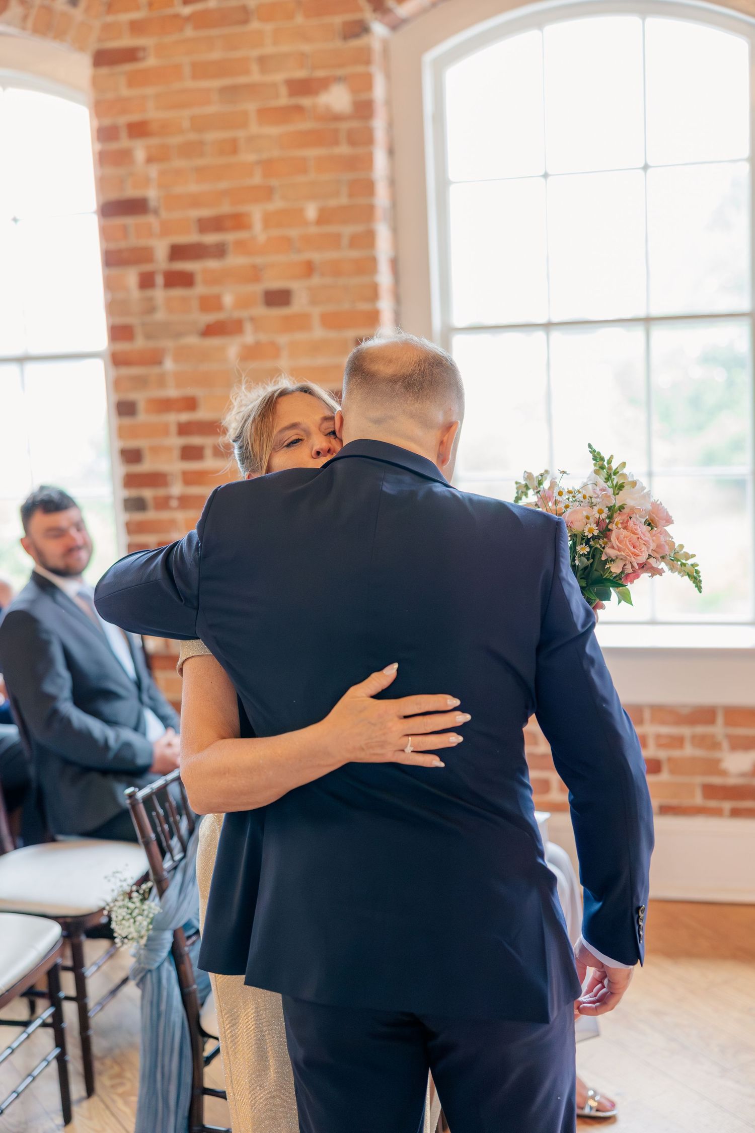 Emotional mother embraces groom in navy suit inside bright brick-walled wedding ceremony venue.