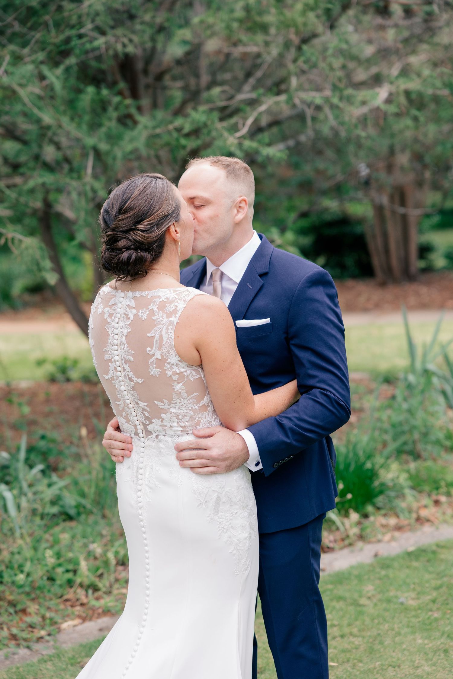 Bride and groom share a romantic kiss outdoors in a lush garden setting, bride wearing elegant lace back wedding gown.