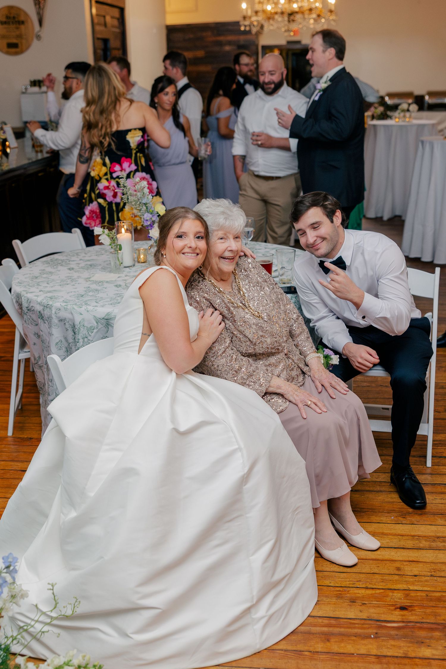 Bride and groom pose with elderly grandmother at wedding reception on wooden floor venue.