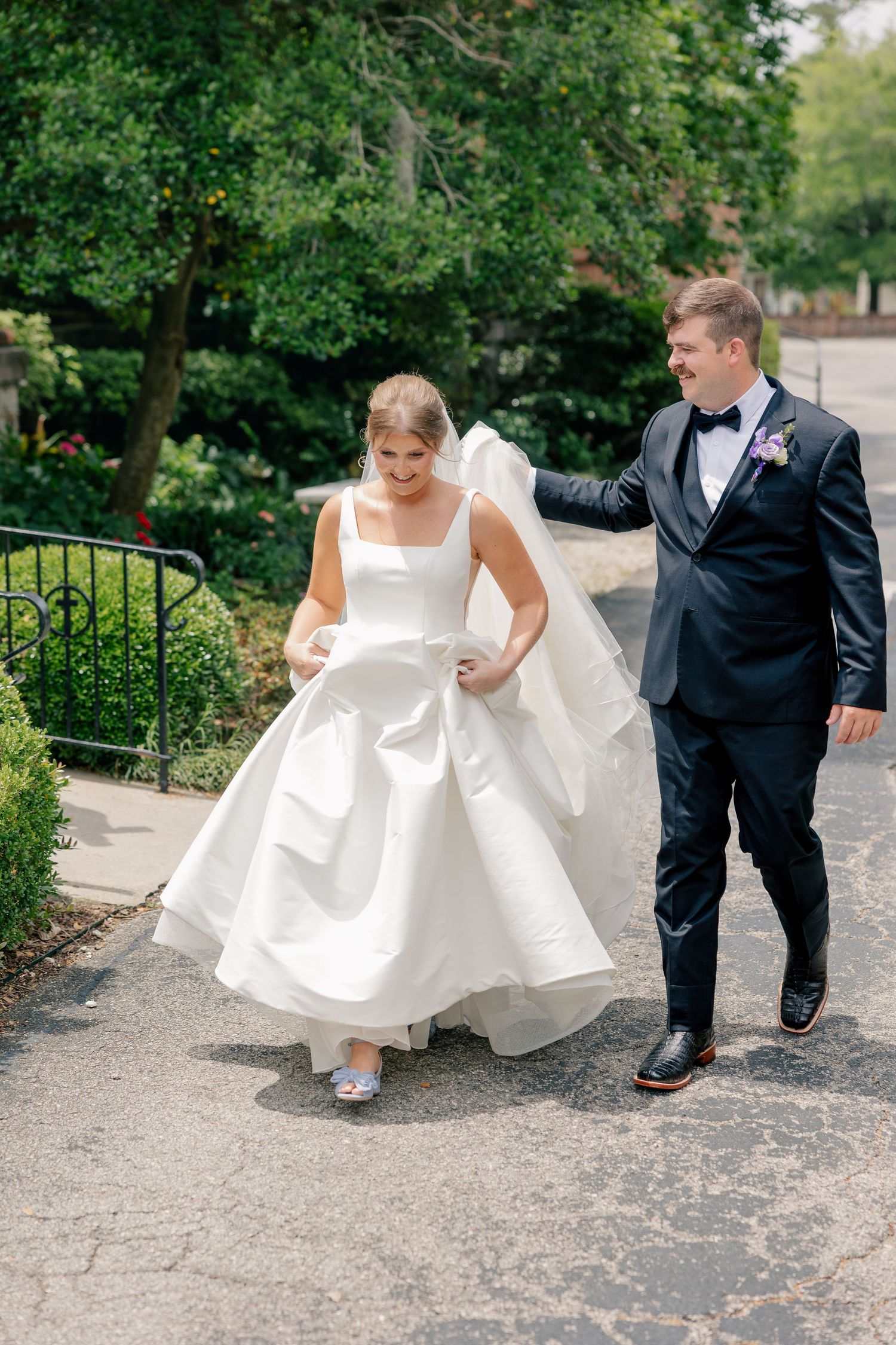 Bride and groom walking outdoors on wedding day, groom in black tuxedo helping carry bride's white ball gown dress.