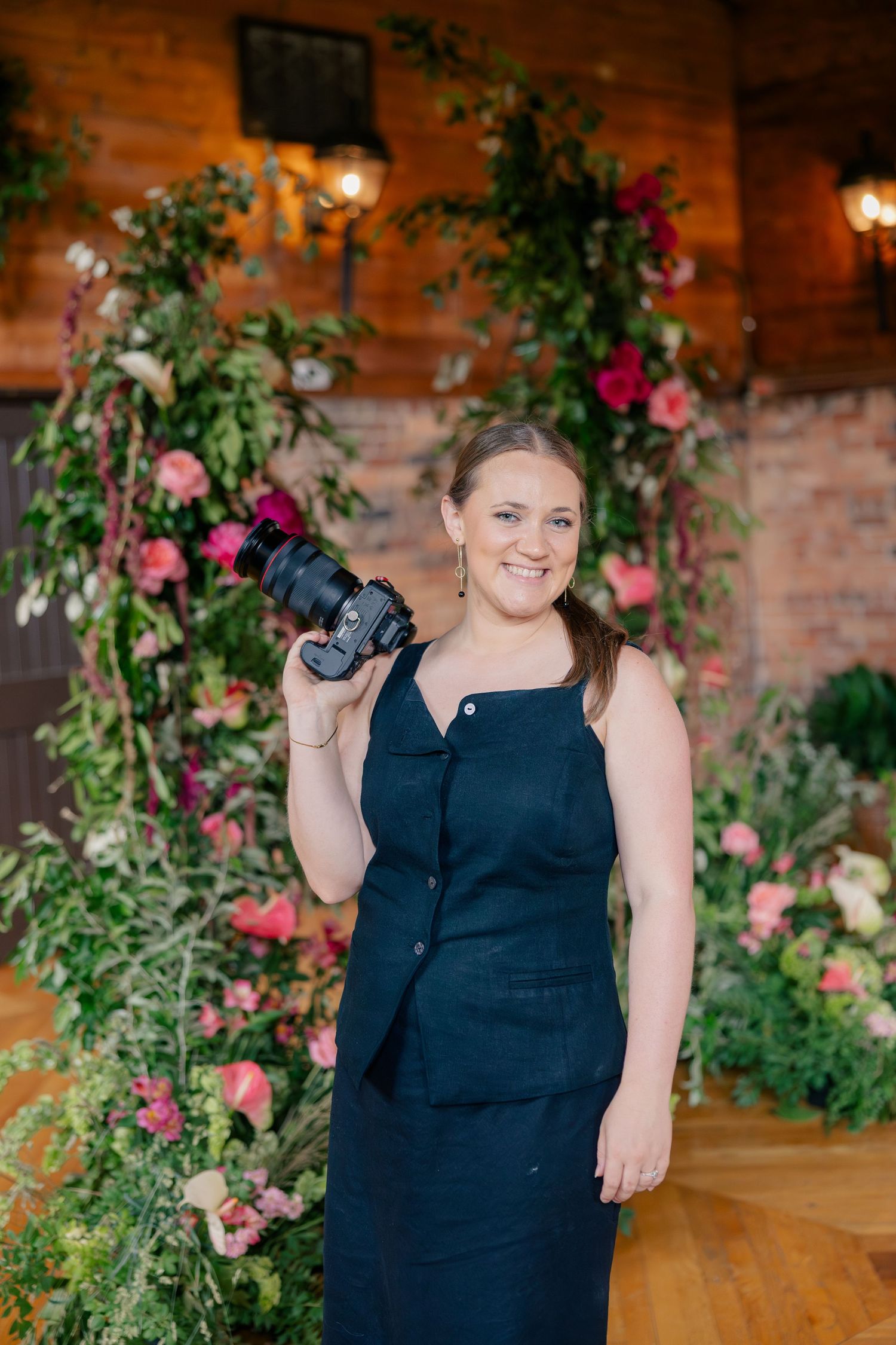 Smiling female photographer in navy dress holding camera, posing in front of floral arch with pink blooms indoors.