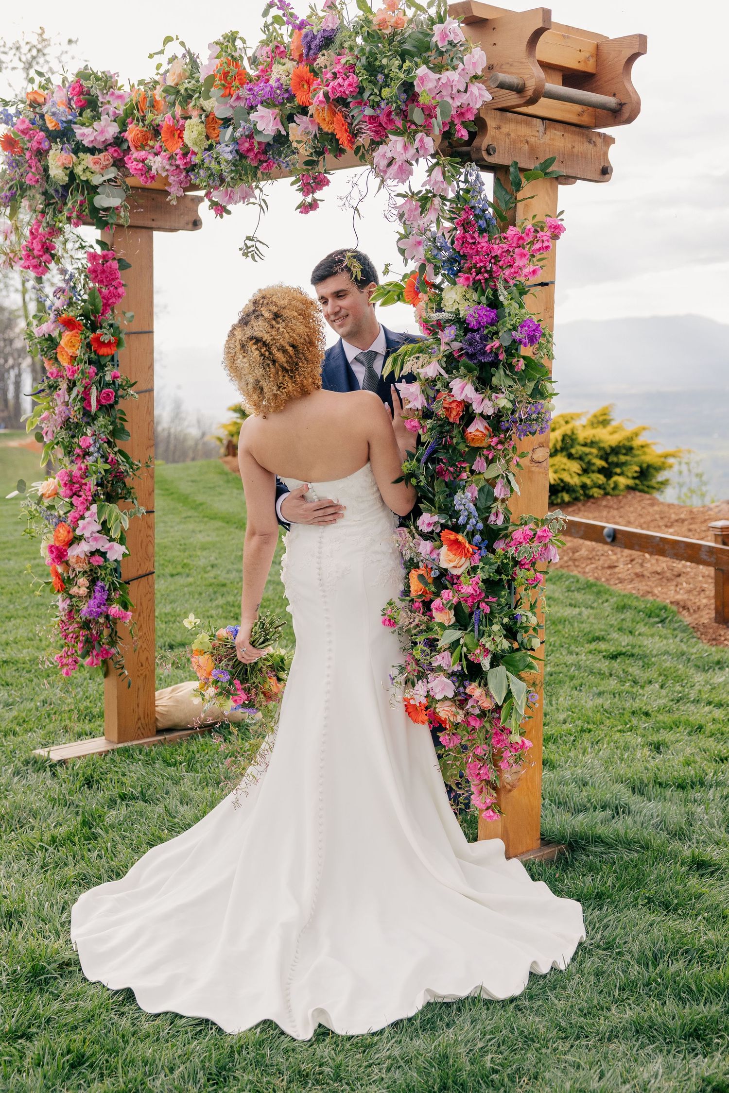 Bride and groom share an intimate moment under a wooden arch adorned with colorful blooms at outdoor wedding.