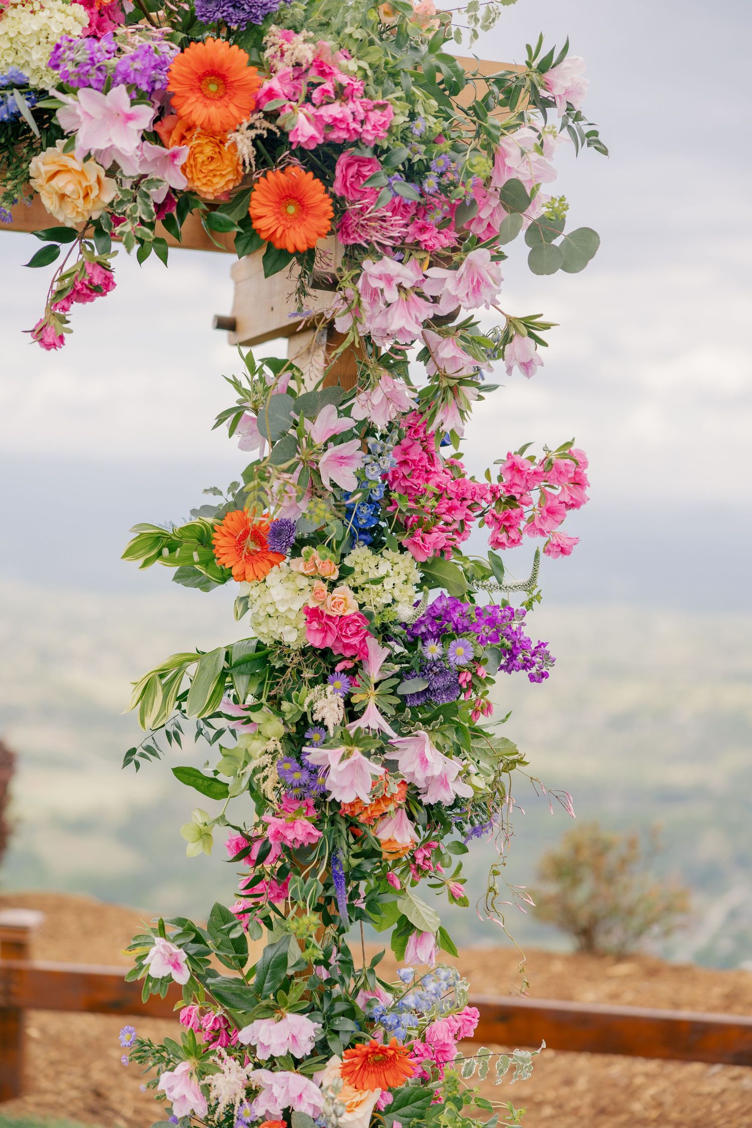 Vibrant colorful floral wedding arch with orange, pink, purple blooms against mountain backdrop.