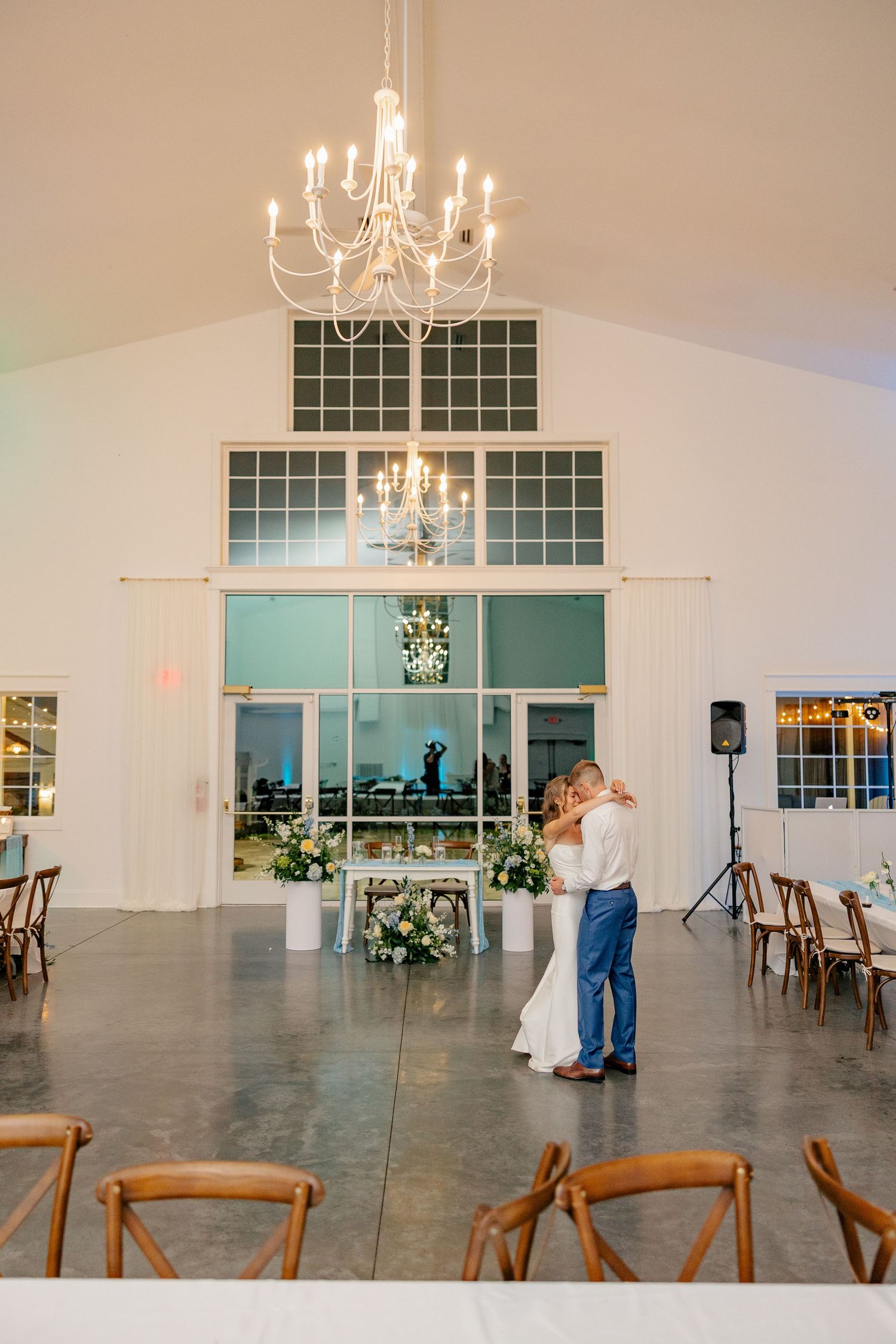 Bride and groom share last dance in white barn venue under chandelier with floral arrangements and wood chairs.