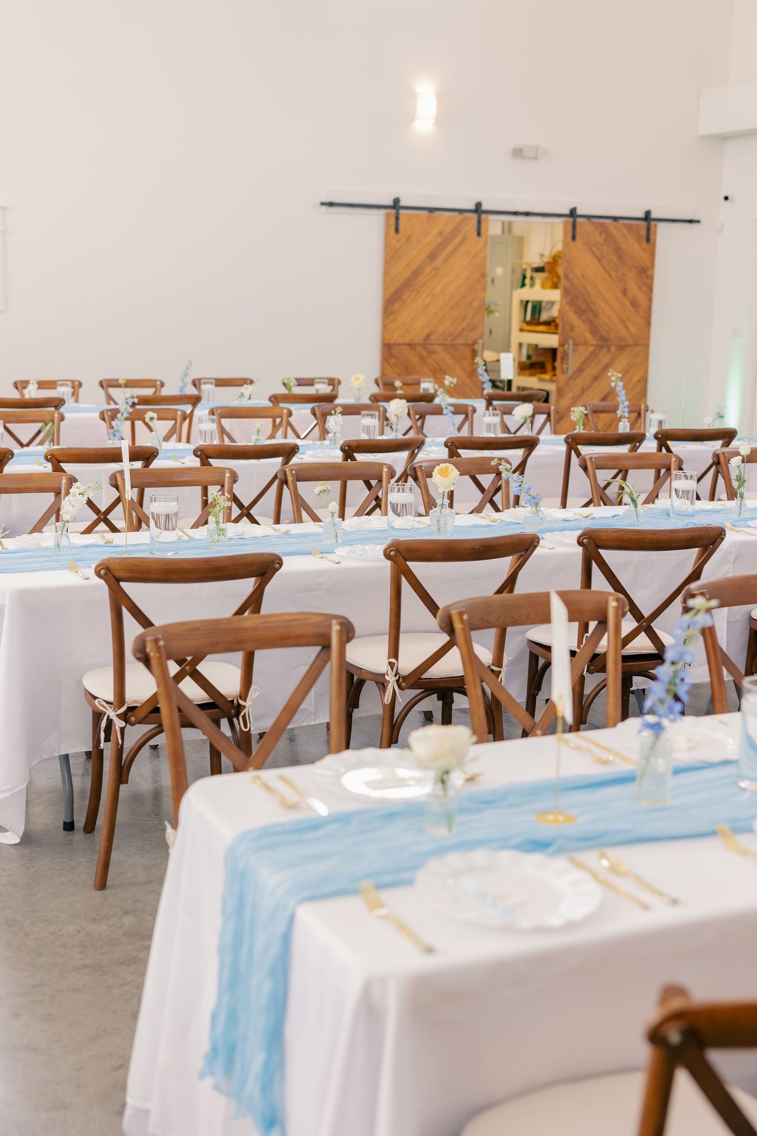 Bright wedding reception hall with wooden cross-back chairs, white tablecloths, and light blue table runners.