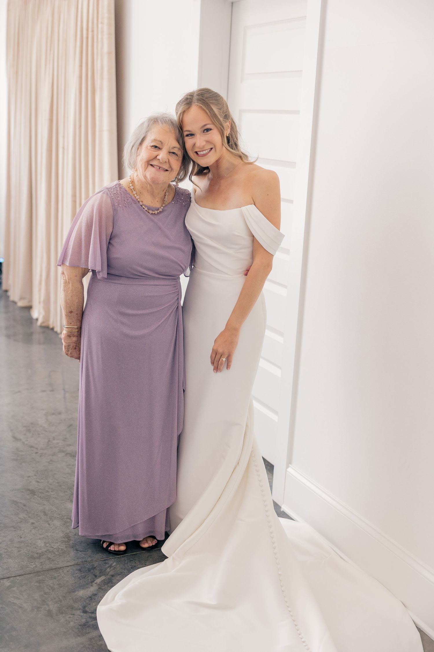 Bride in white off-shoulder gown poses with older woman in lavender dress at wedding venue.