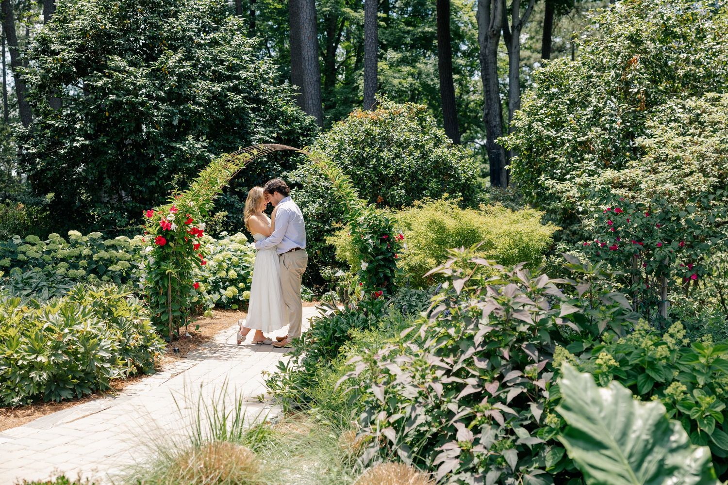 Couple embracing under floral garden arch surrounded by lush green foliage on a brick pathway.