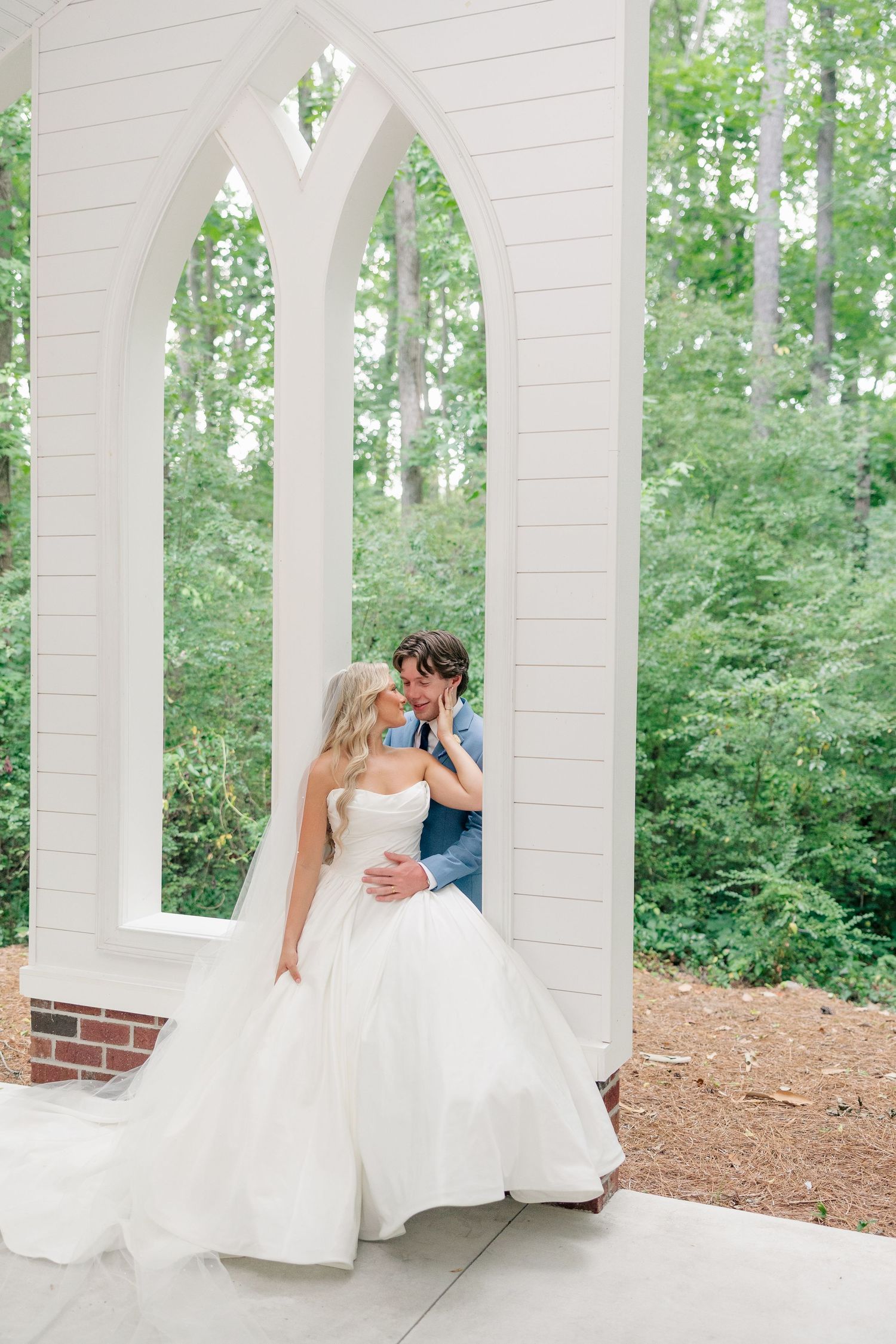 Bride and groom share a romantic kiss under white Gothic arched windows surrounded by lush greenery.