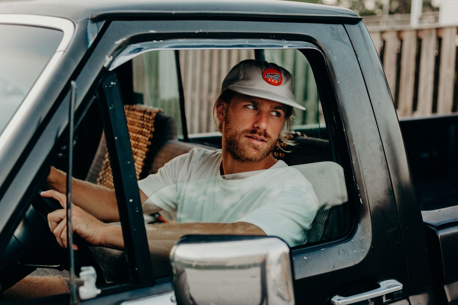 Someone sitting in the driver's seat of a truck wearing a baseball cap and white t-shirt in natural lighting.