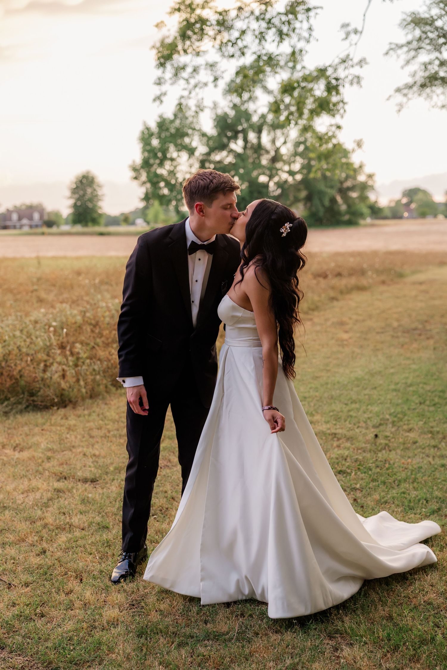 A romantic sunset wedding photo of a couple in formal attire standing in a grassy field.