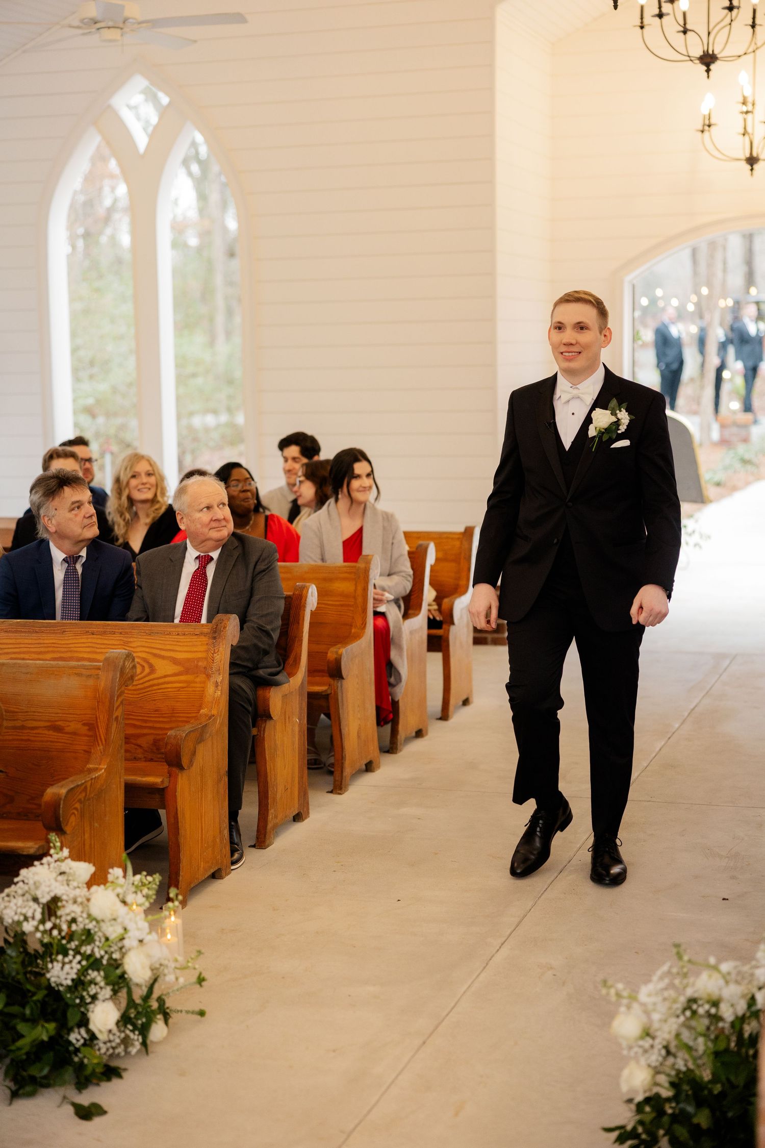 Wedding ceremony in a white chapel with wooden pews decorated with floral arrangements.