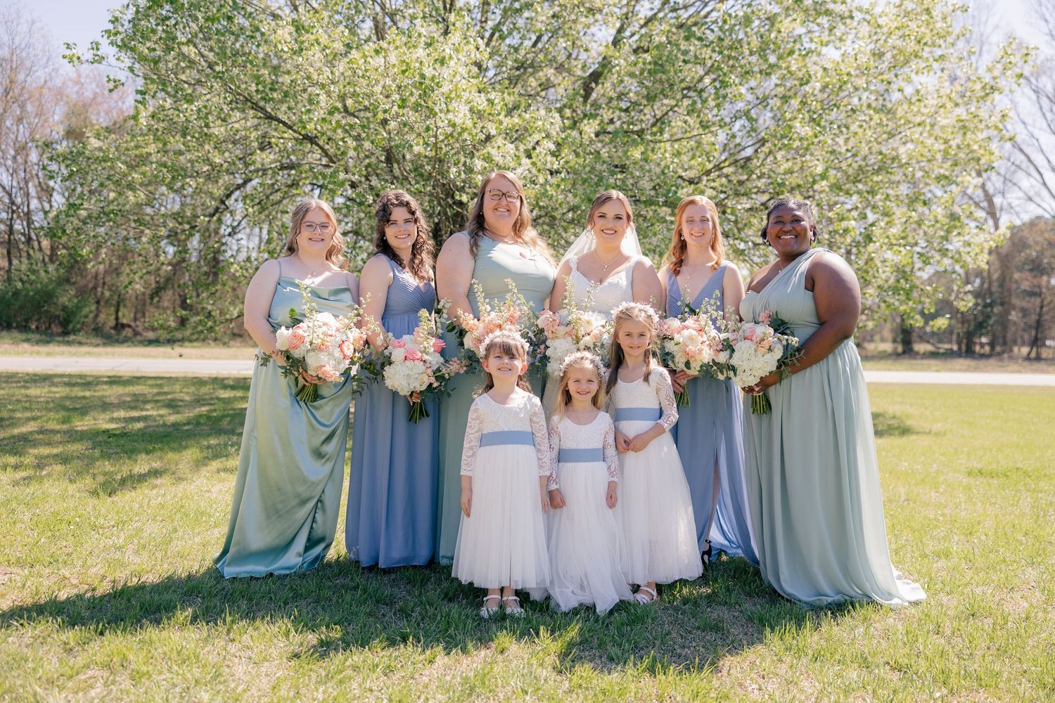 Bridesmaids wearing different shades of blue and mint dresses pose outdoors with flower girls in white dresses.