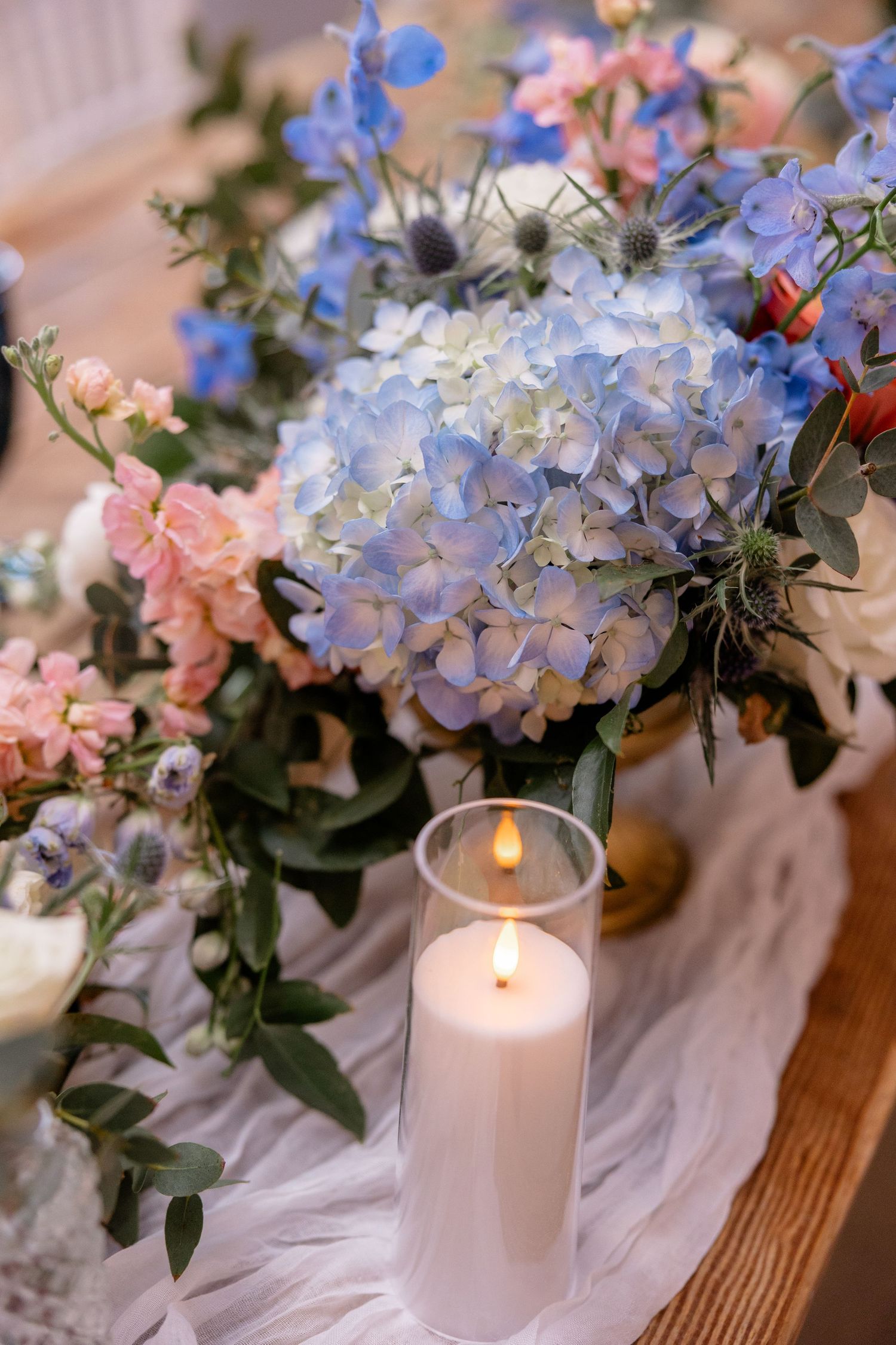 Table centerpiece with blue hydrangeas, pink roses and white pillar candles creating soft ambiance.