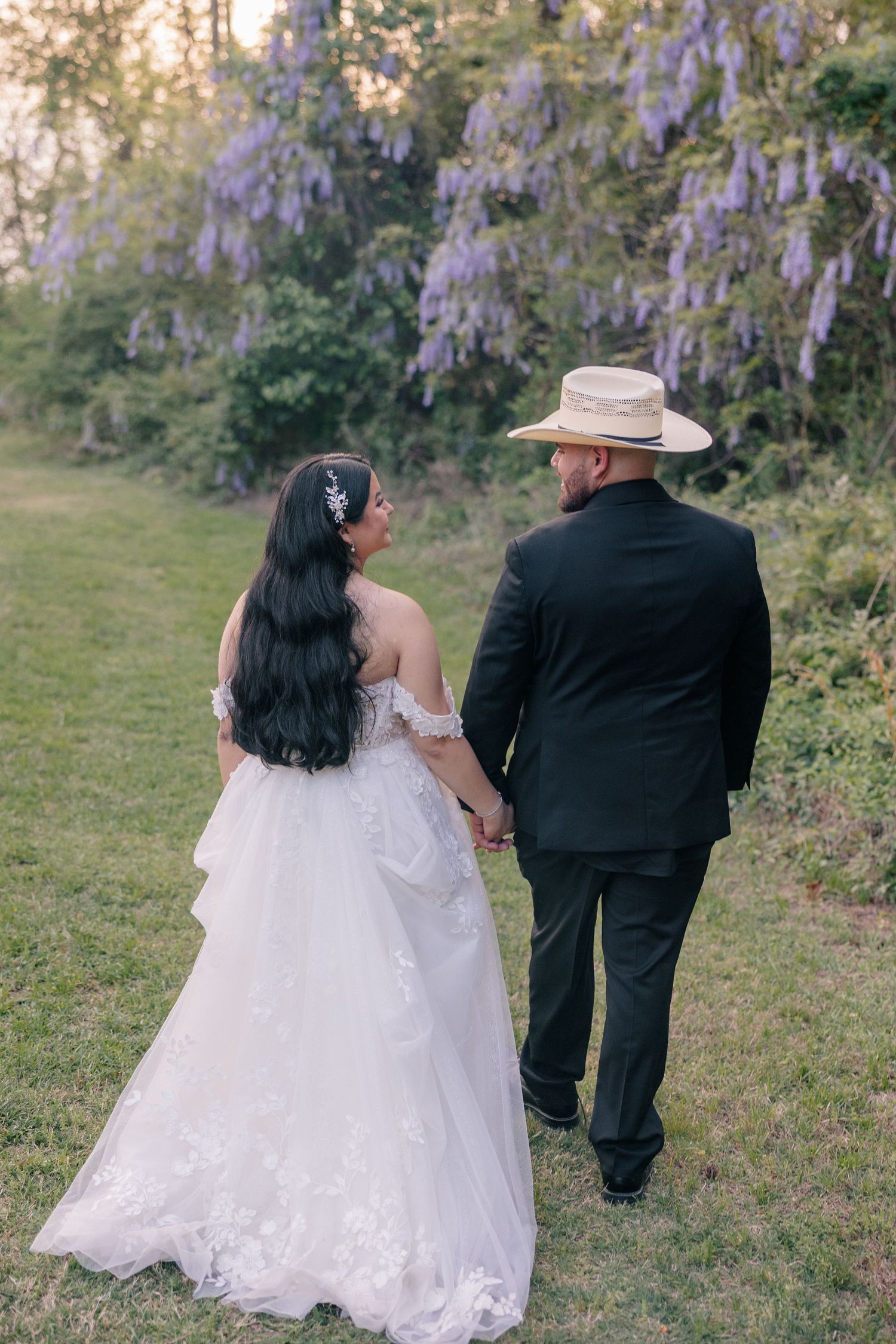 A couple walking through a garden with wisteria, bride in white gown and groom wearing a cowboy hat.