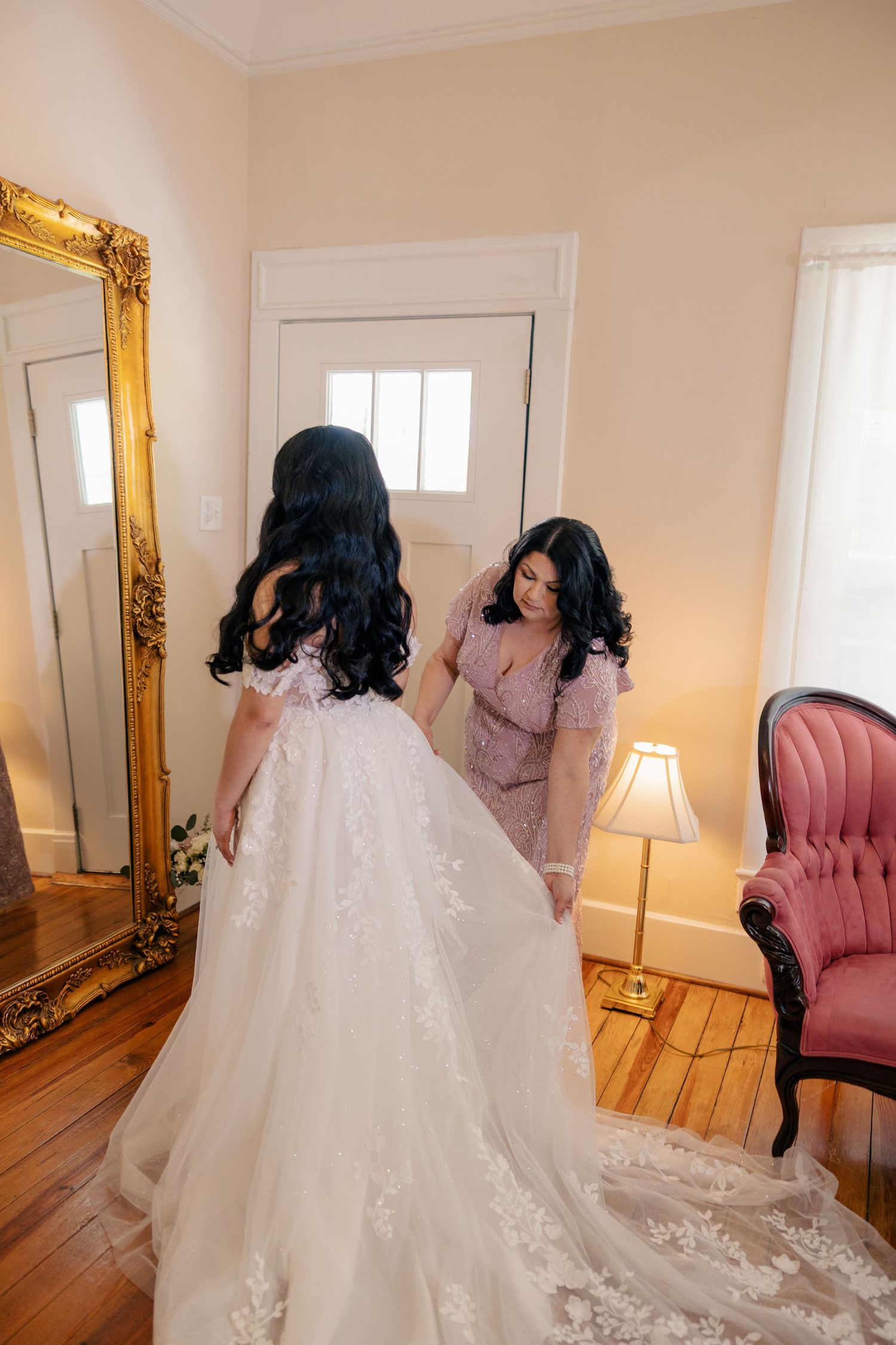 White wedding gown with lace details displayed in an elegant room with pink velvet chair and ornate mirror.