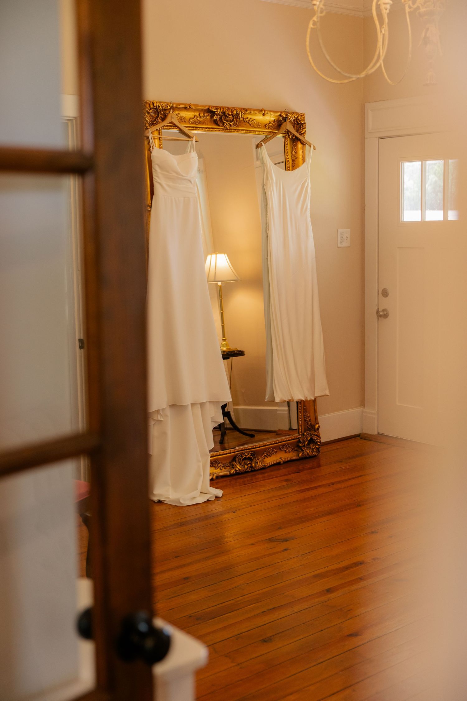 Two elegant white wedding dresses hang on a gilded ornate mirror in a room with warm lighting and hardwood floors.