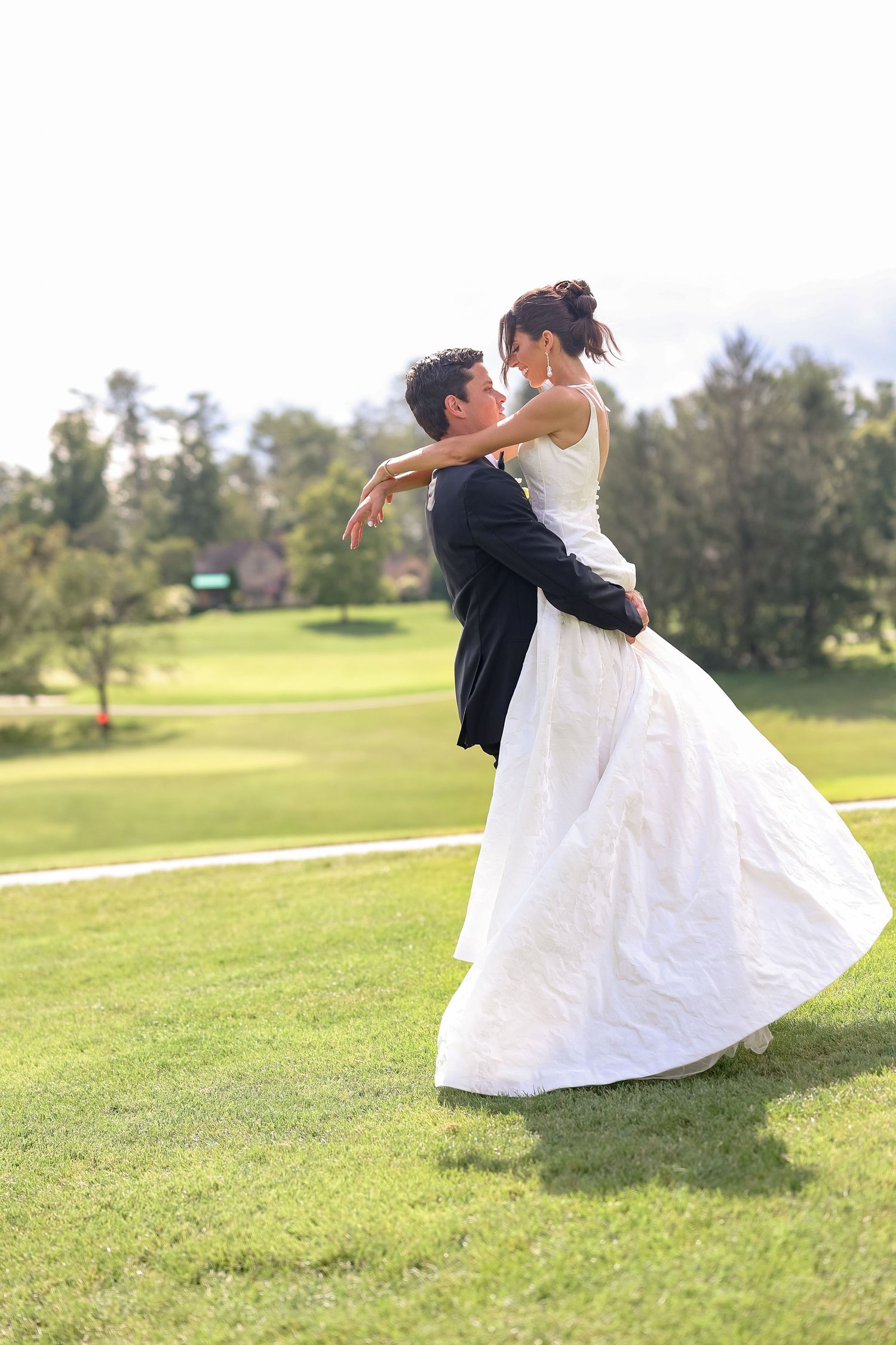 Groom lifts bride in white wedding dress on lush green lawn during joyful outdoor wedding celebration.